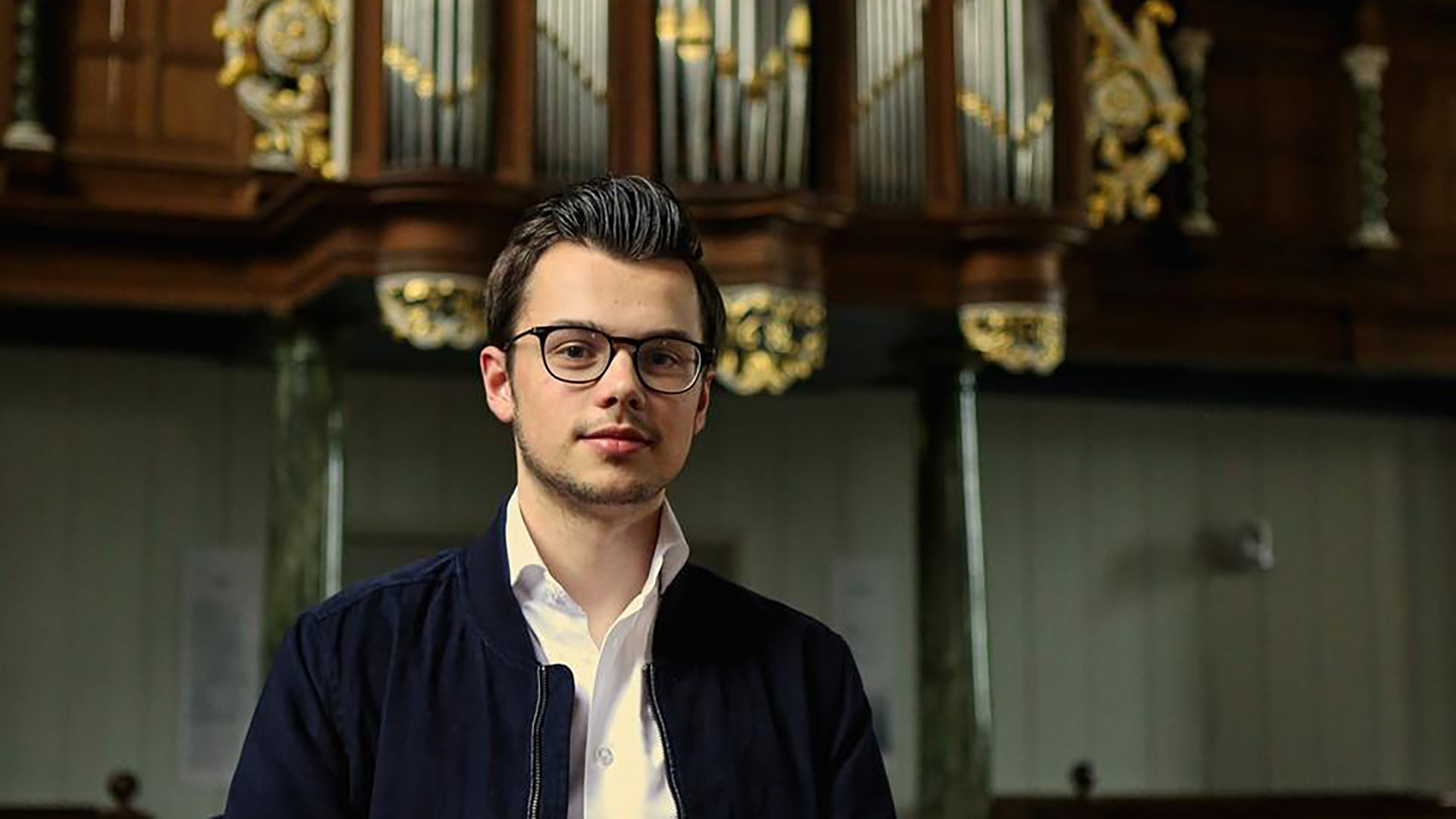 Een man met een bril en donker haar staat voor een decoratief kerkorgel in een kerkelijke setting.