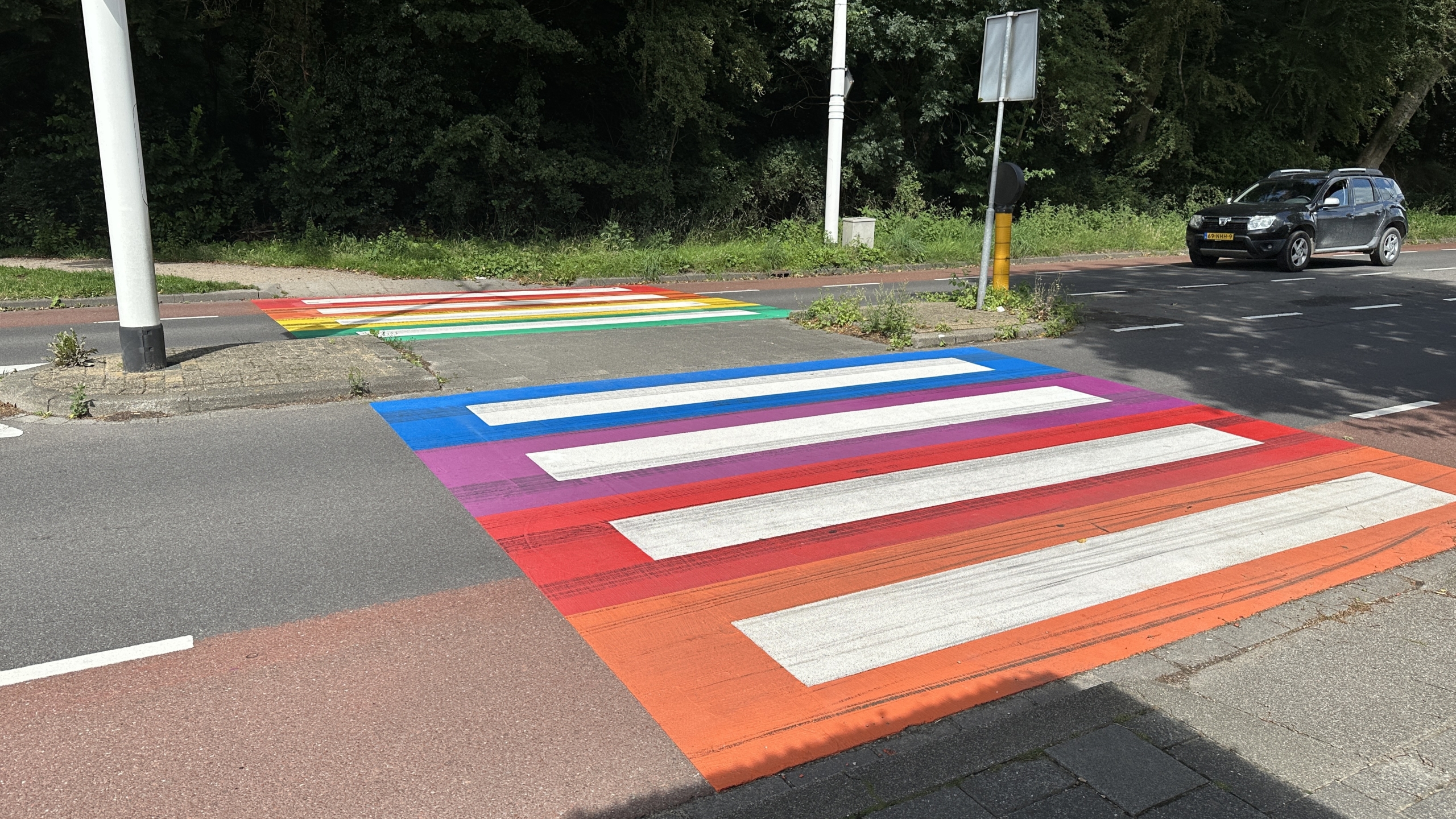 Een zebrapad in regenboogkleuren in een straat met een voorbijrijdende auto en bomen op de achtergrond.