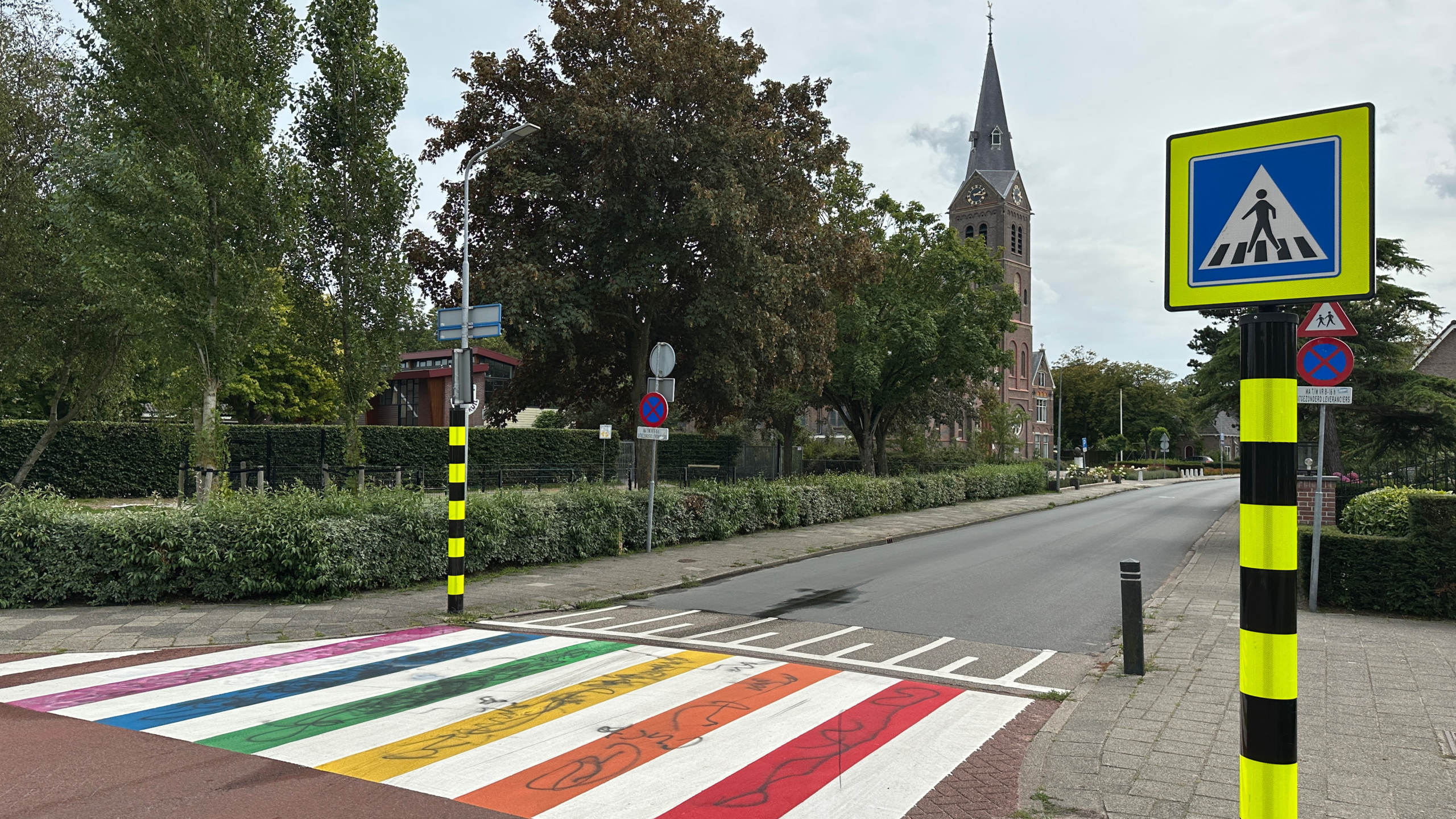 Straatbeeld met een regenboogzebrapad en verkeersborden in een groene omgeving, met een kerk op de achtergrond.