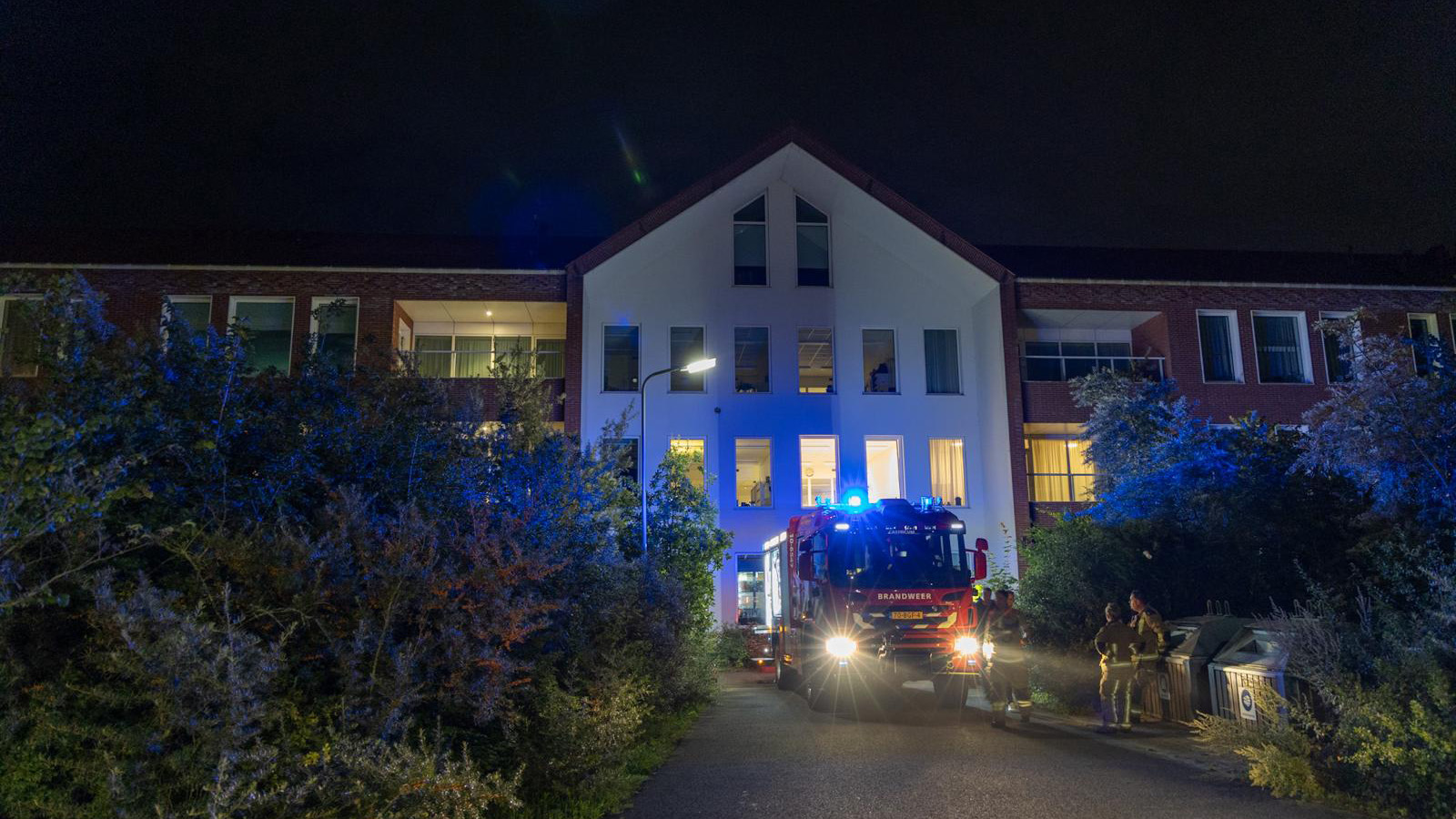 Een brandweerwagen met knipperende lichten staat 's nachts voor een groot gebouw met meerdere verdiepingen, omgeven door struiken. Brandweerlieden staan naast de wagen.