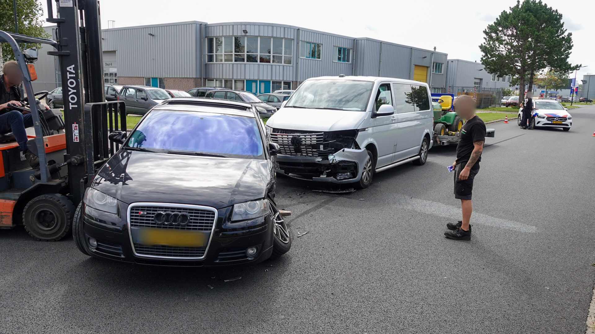 Een zwarte auto en een witte bestelwagen zijn beschadigd tijdens een botsing op een industrieel terrein. Een vorkheftruck helpt bij het verplaatsen van de zwarte auto. Een man staat naast de voertuigen terwijl een politieauto op de achtergrond staat.