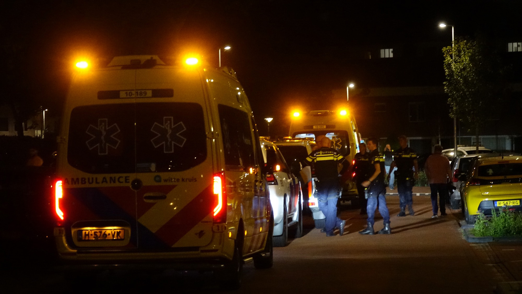 Ambulance en politievoertuigen staan geparkeerd langs een straat 's nachts, met brandende waarschuwingslichten en politieagenten die bij de voertuigen staan.
