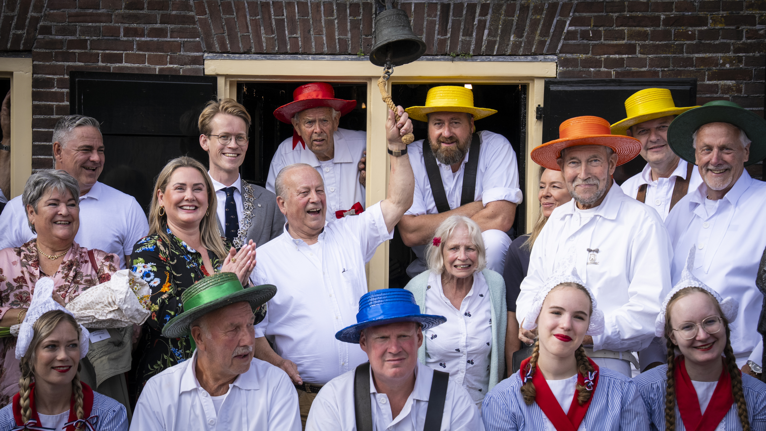 Groep mensen in traditionele Nederlandse kleding poseren voor een bakstenen gebouw, met een man die een bel luidt; de hoeden in verschillende kleuren geven de sfeer van een feestelijke gelegenheid weer.