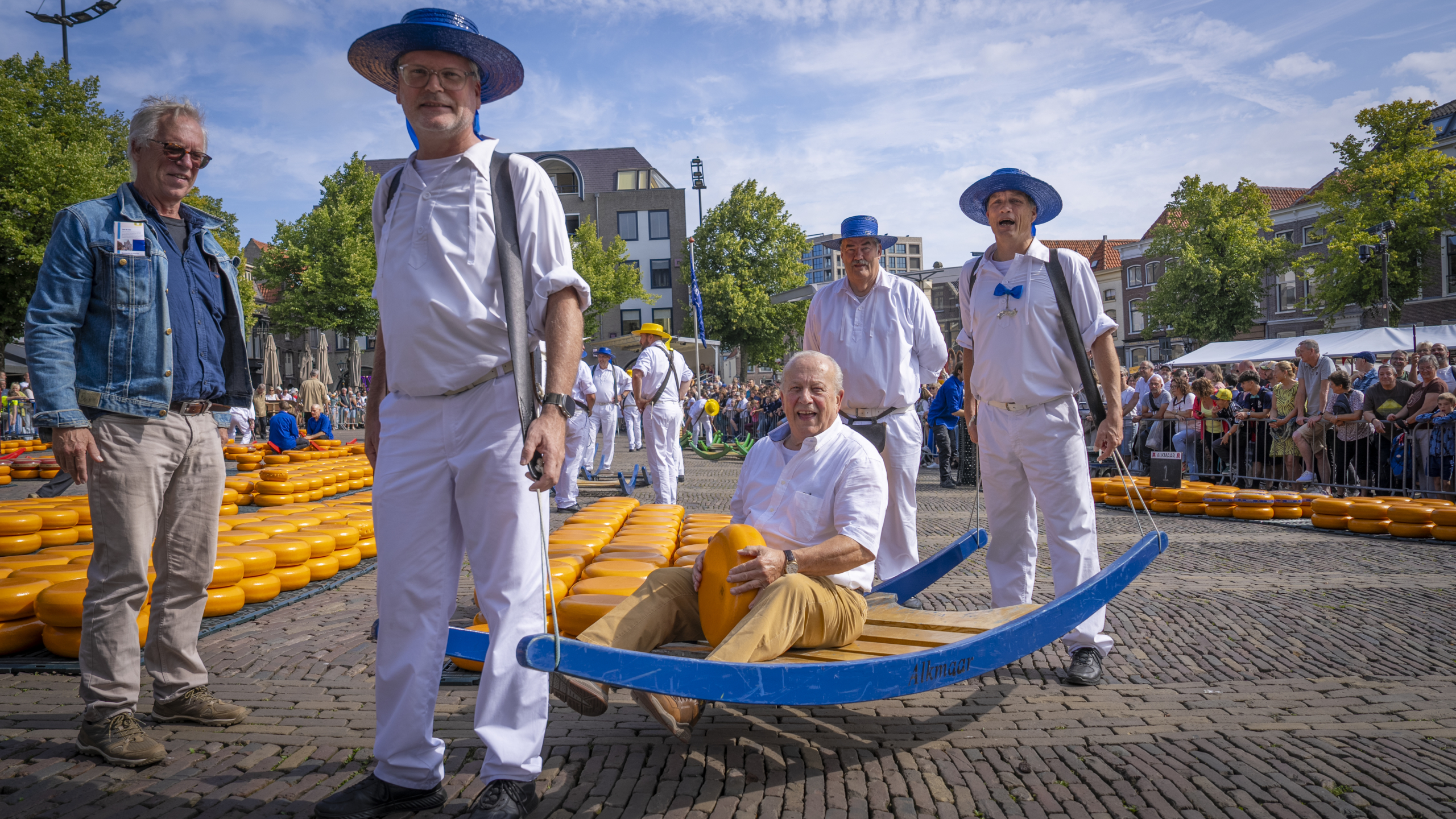 Mannen in witte kleding en blauwe hoeden dragen een kaasbrancard met ronde kazen op een marktplein, omringd door toeschouwers en gebouwen.