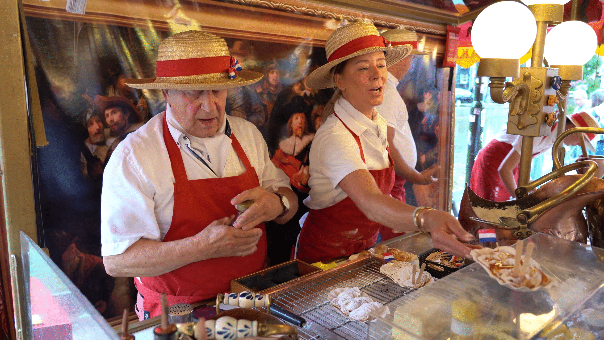 Twee personen met strohoeden en rode schorten werken bij een eetkraampje, met wafels en ingrediënten voor zich, tegen een achtergrond van een klassiek schilderij.