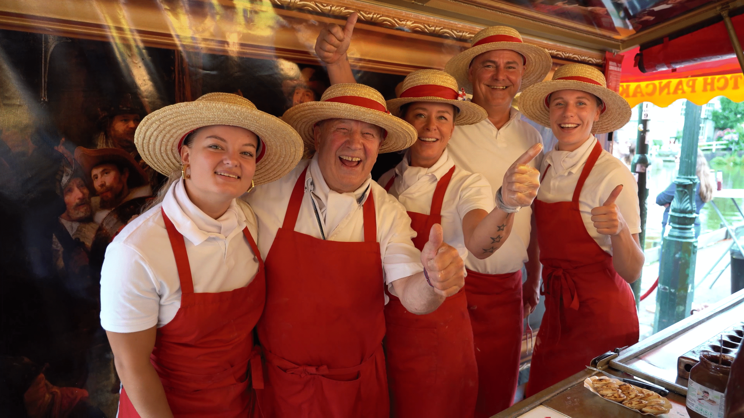 Vijf lachende mensen, gekleed in witte shirts en rode schorten, dragen strohoeden en staan in een rij terwijl ze hun duimen omhoog steken in een kraam.