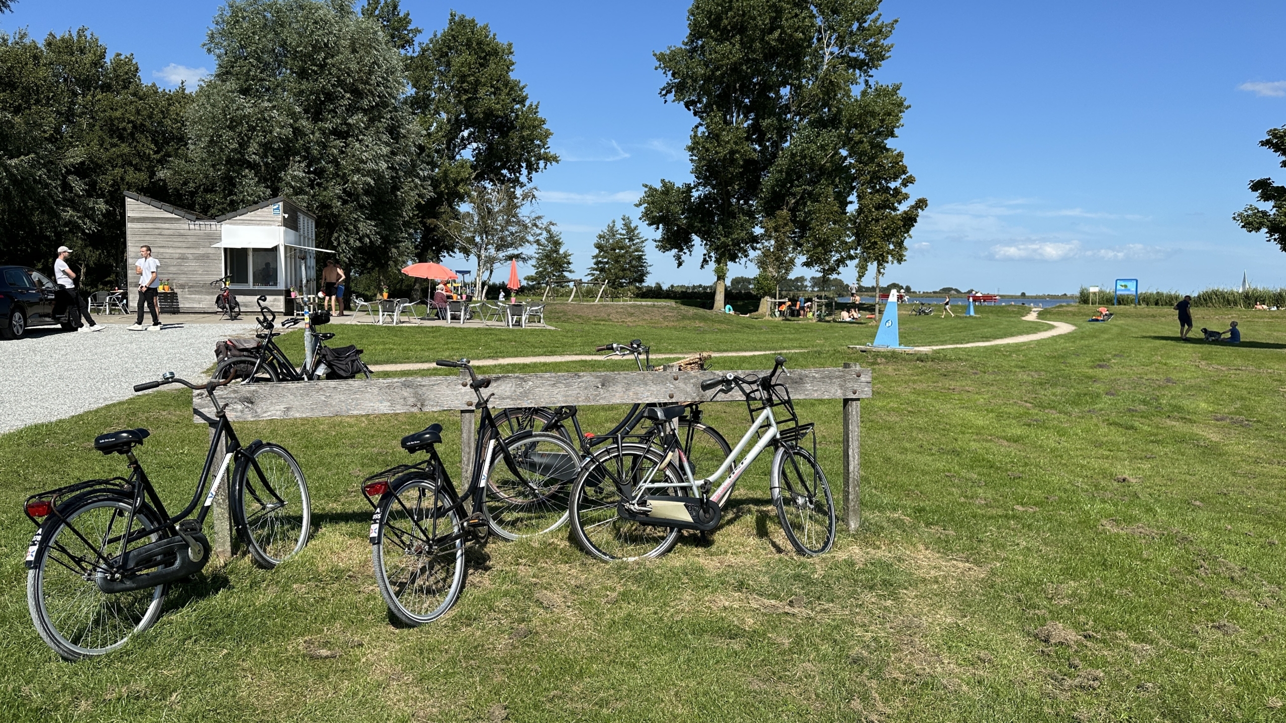 Fietsen staan geparkeerd bij een houten fietsenrek op een grasveld met een pad dat naar een meer leidt; mensen zitten en wandelen rond een verkoopkiosk. Er zijn enkele bomen en een duidelijk blauwe hemel op de achtergrond.