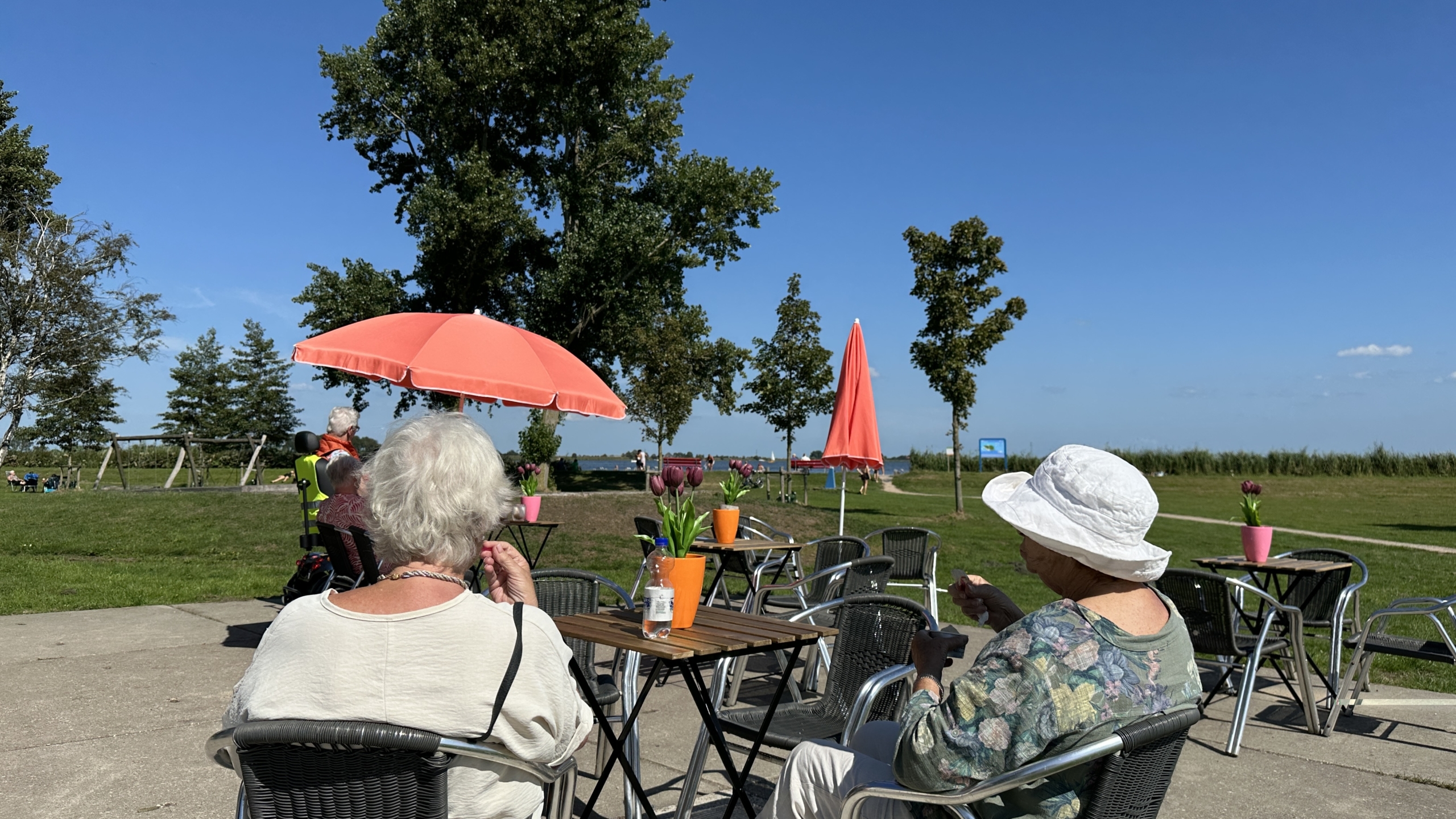 Twee oudere vrouwen zitten op een terras met tafels en stoelen, onder een roze parasol op een zonnige dag, met uitzicht op een groene omgeving en blauwe lucht.