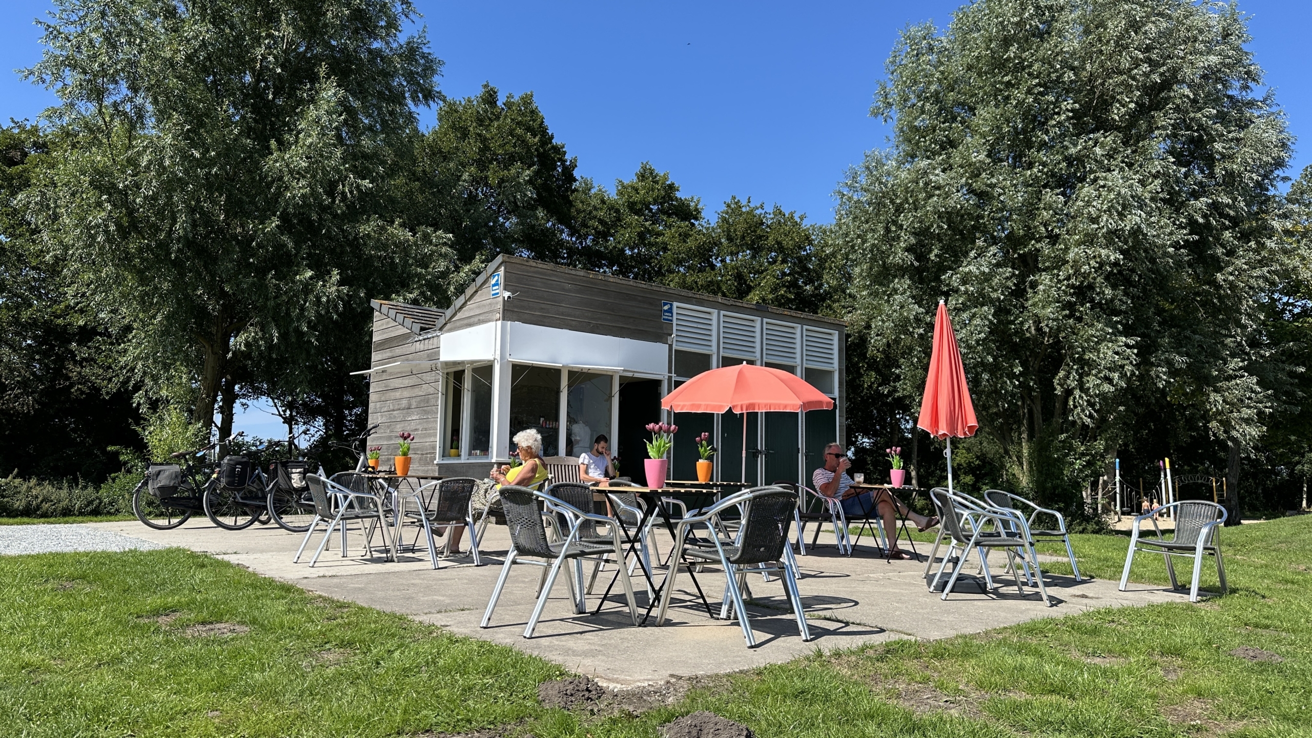 Een klein café met een terras met enkele tafels en stoelen onder rode parasols, waar mensen genieten van drankjes in een groene omgeving met grote bomen.