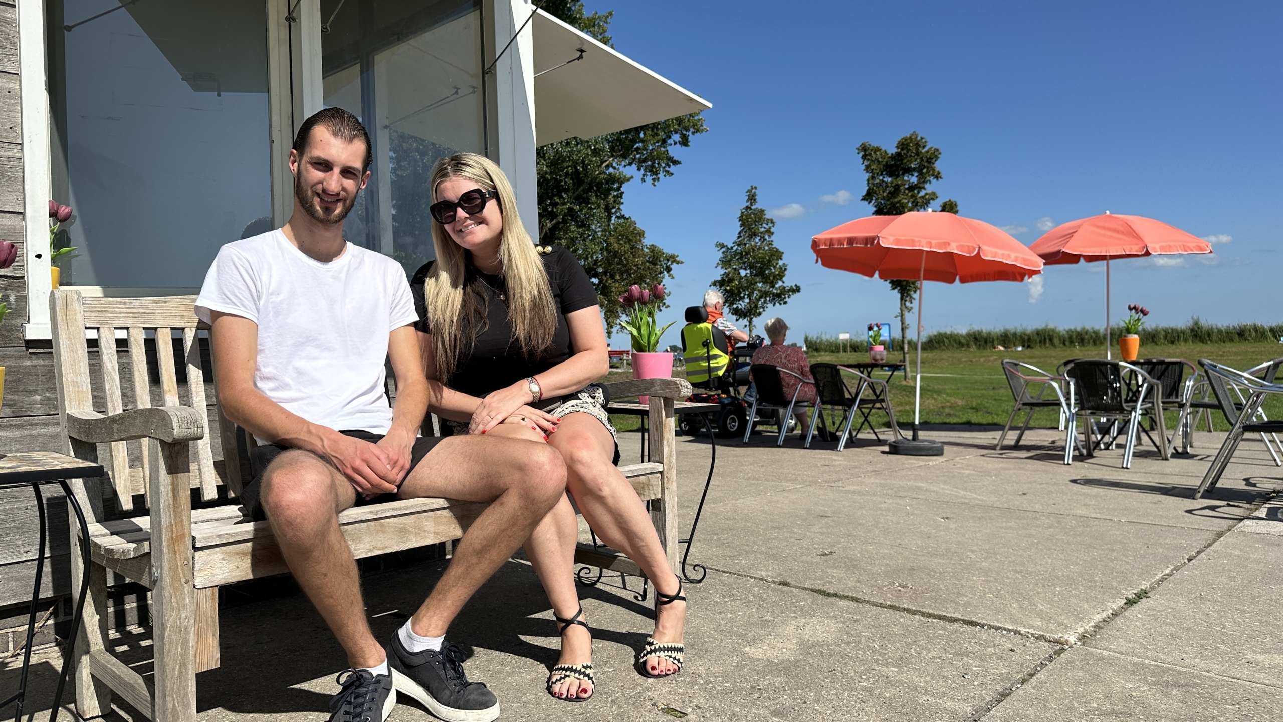 Een man en een vrouw zitten op een houten bankje buiten, met een heldere blauwe lucht en rode parasols op de achtergrond. Ze glimlachen en genieten van de zon, omgeven door tafels en stoelen van een terras.