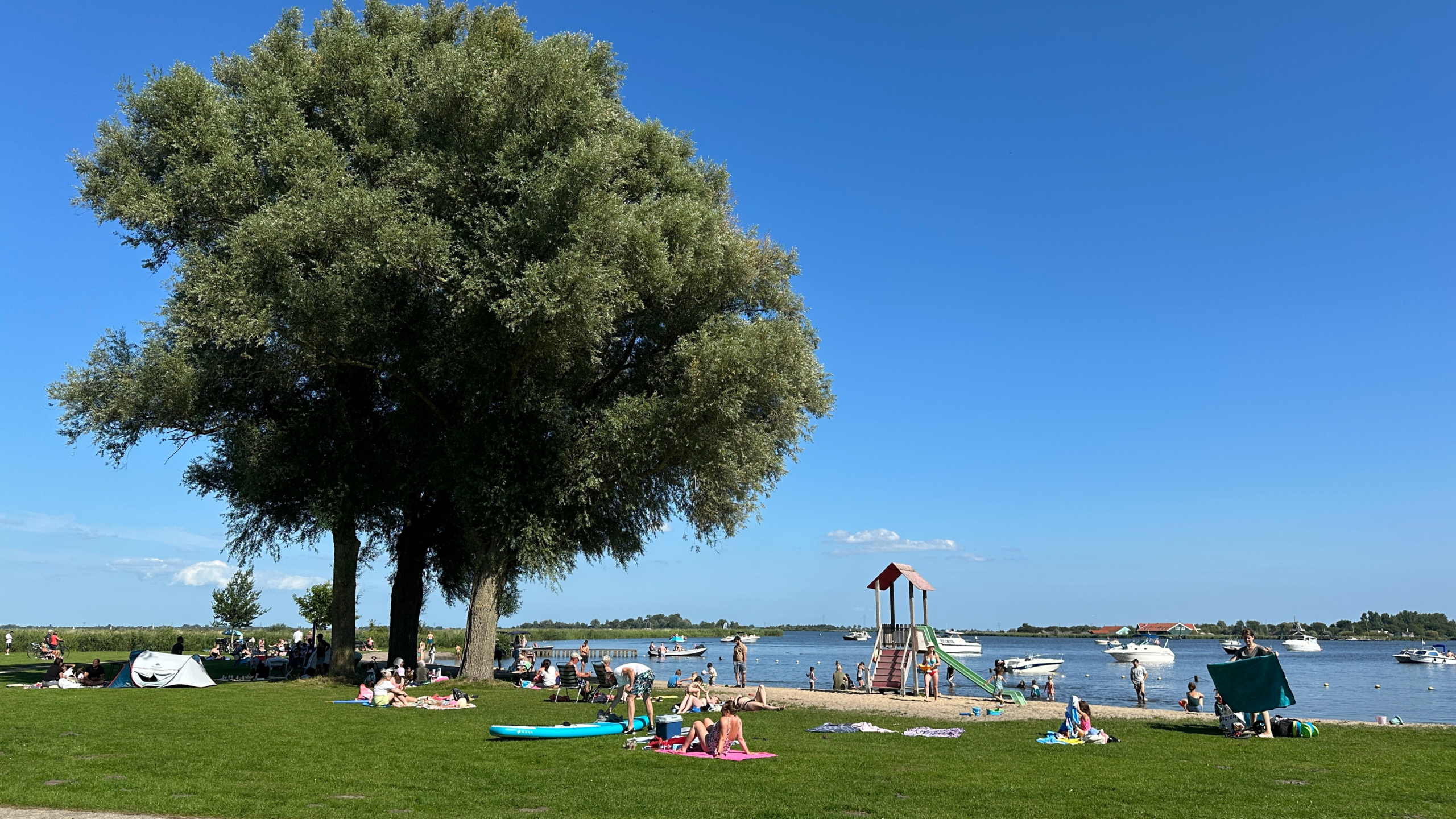 Mensen ontspannen op een grasveld naast een meer met enkele boten en een speeltoestel in de buurt, onder een heldere blauwe hemel.