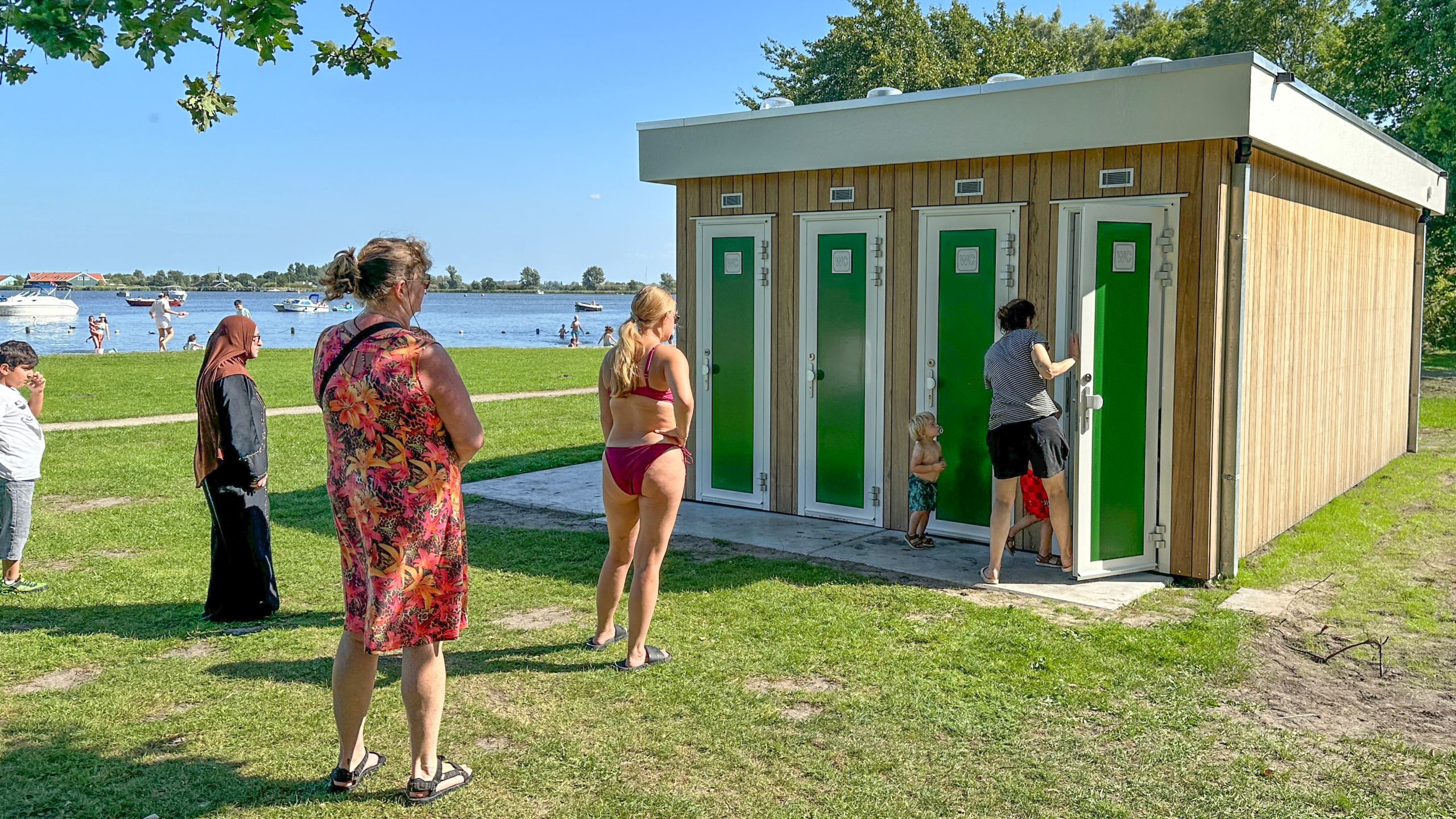 Mensen staan in de rij voor een gebouw met houten gevel en groene deuren op een strandgrasterrein, met in de achtergrond een meer en zwemende mensen.