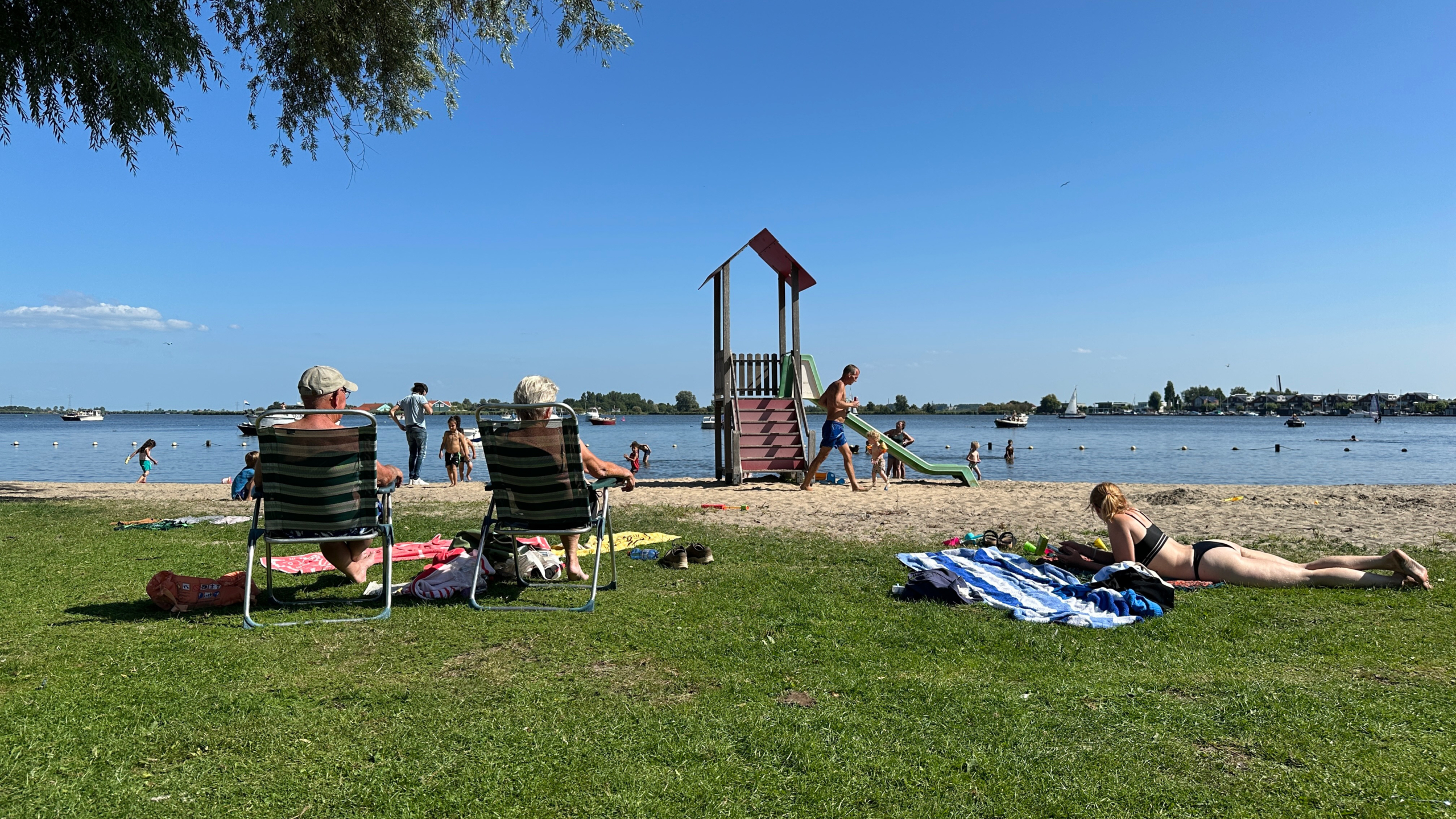 Mensen ontspannen op een grasveld bij een strand met uitzicht op een rustig meer op een zonnige dag. Een speeltoestel met glijbaan staat op het zandstrand, terwijl kinderen in het water spelen en volwassenen op ligstoelen zitten of op handdoeken liggen.