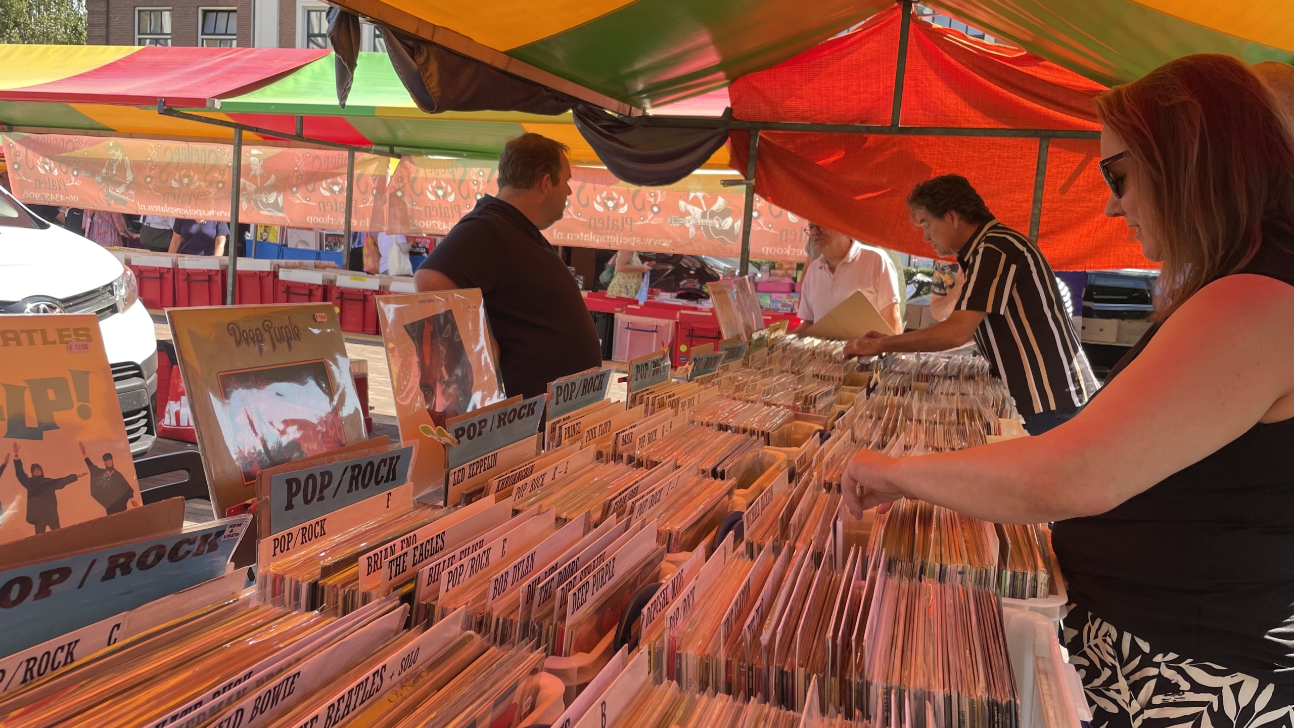 Mensen bladeren door platen in bakken onder een kleurrijke marktkraam, met LP's van artiesten zoals The Beatles, Deep Purple en Led Zeppelin zichtbaar.