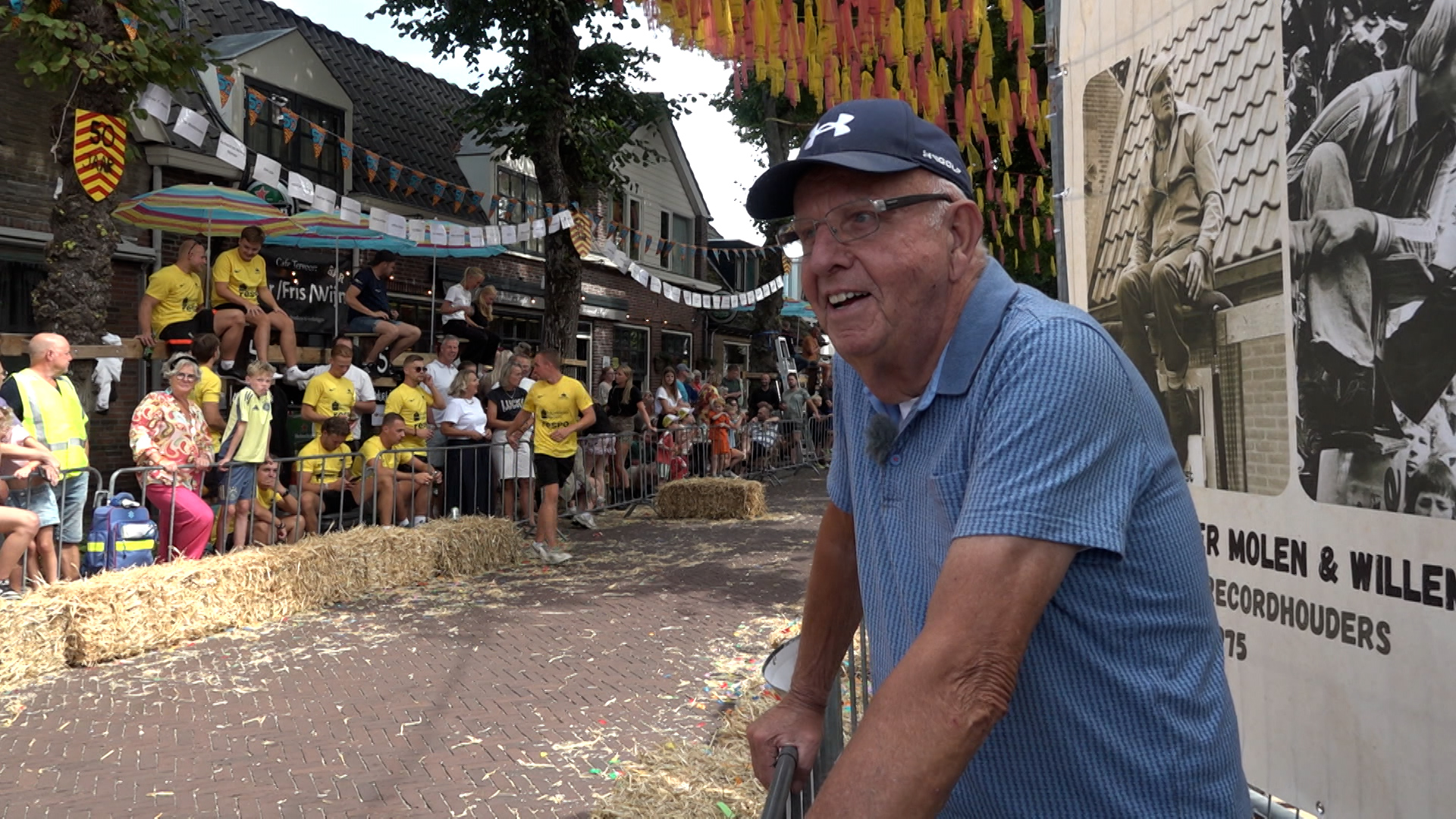 Een oudere man met een blauwe pet en hemd leunt tegen een hek tijdens een evenement. Op de achtergrond zijn toeschouwers te zien bij een straatversiering met strobalen, vlaggen en een groot zwart-wit poster.