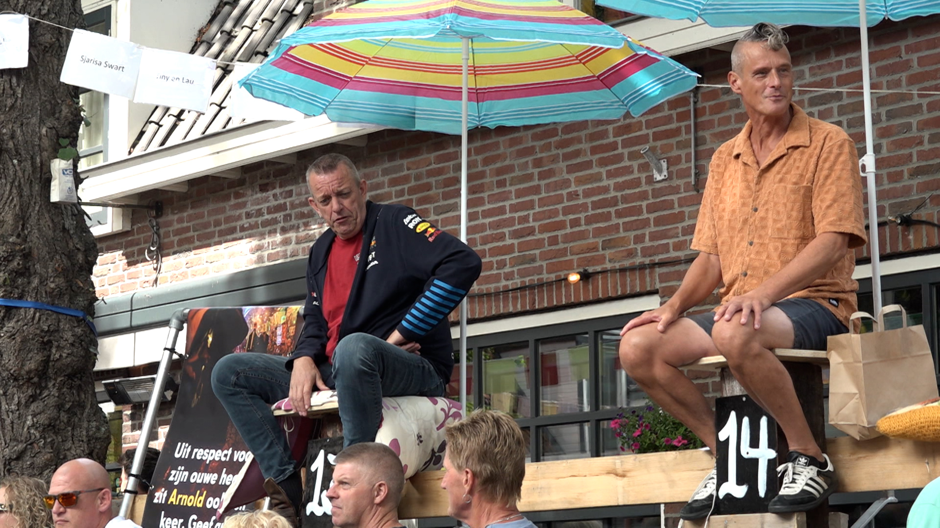 Twee mannen zitten op houten platforms met nummers, beschermd door gekleurde parasols voor een gebouw met bakstenen muur en ramen, omringd door enkele toeschouwers.