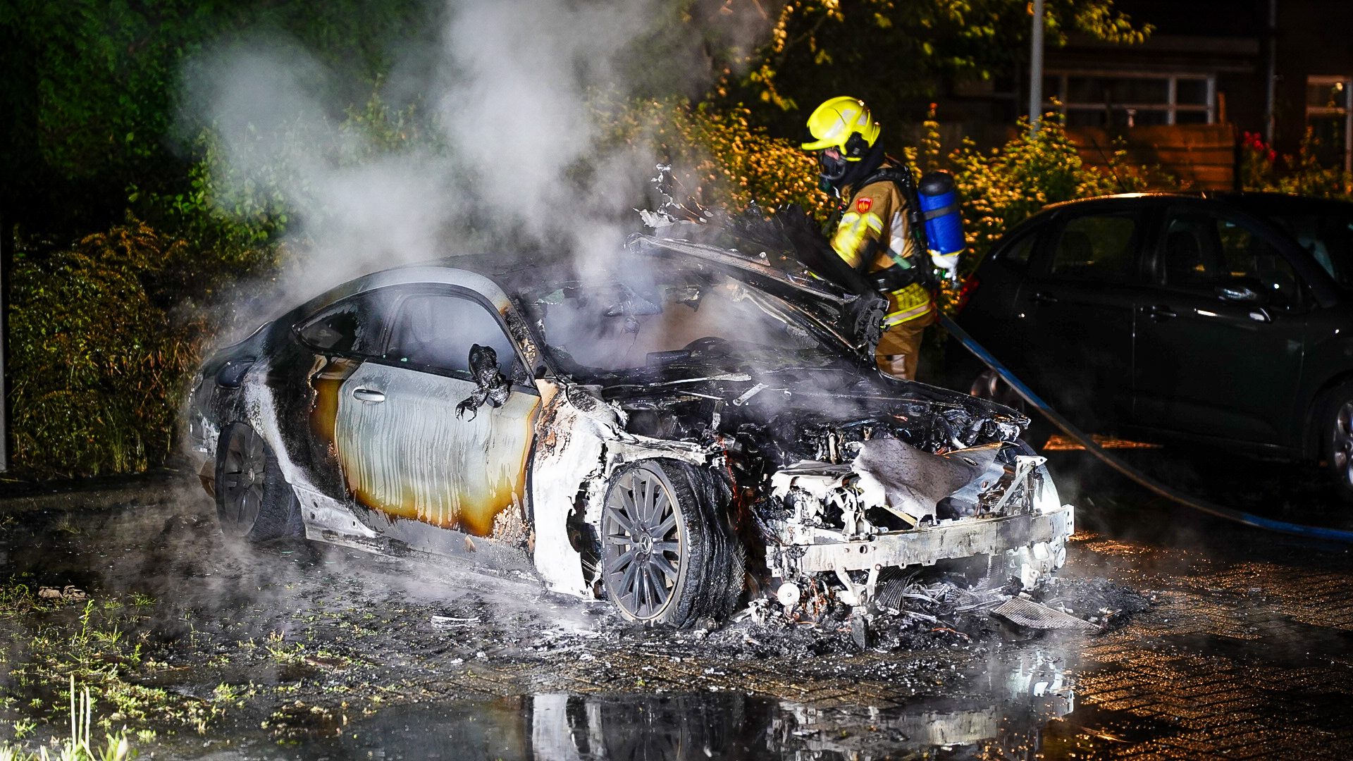 Een brandweerman blust een uitgebrande auto die zwaar beschadigd is door een nachtelijke brand, met rook die nog steeds opstijgt van het voertuig.