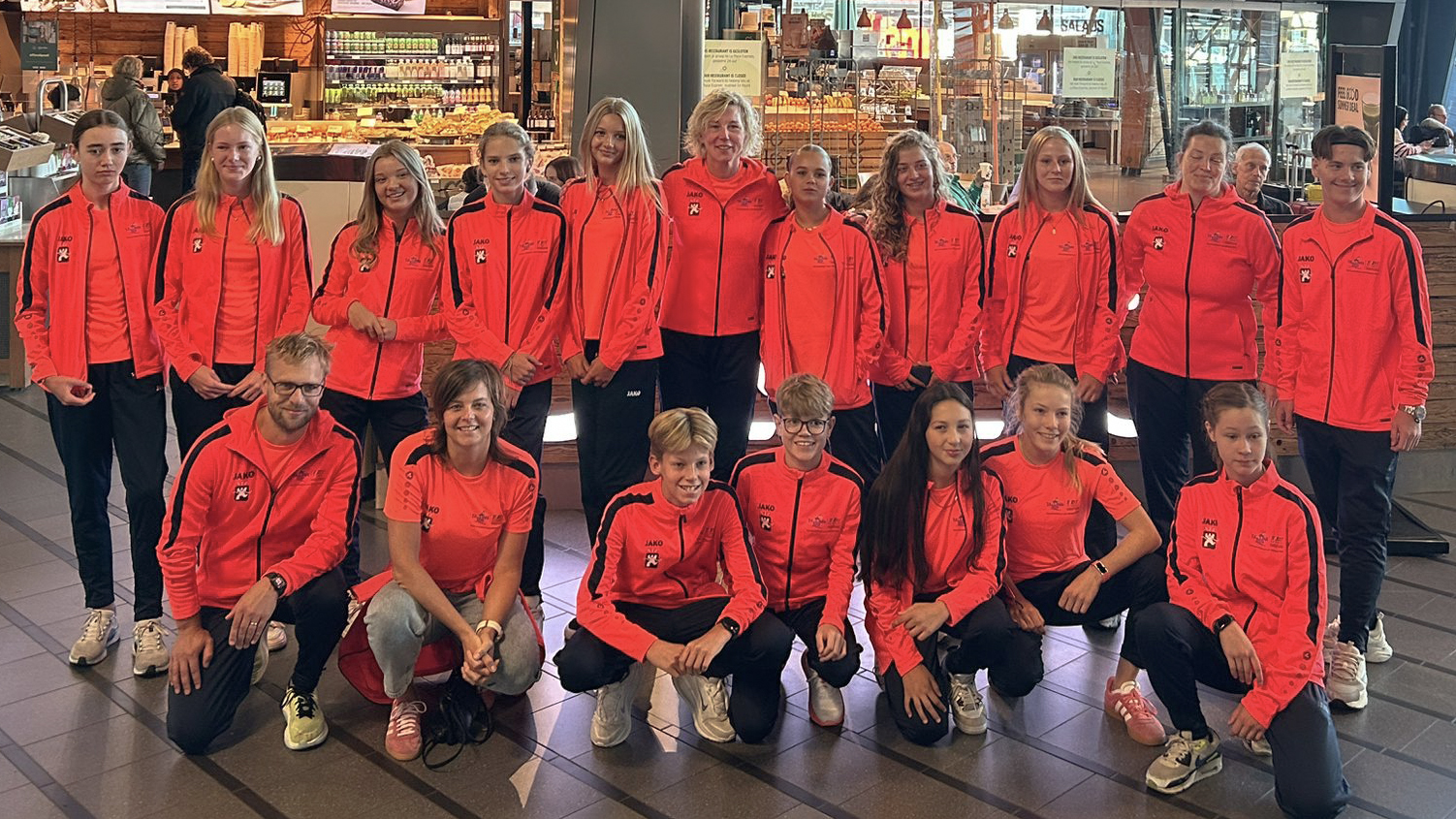 Een groep mensen draagt feloranje sportjacks met een Jako-logo en poseert in een winkelcentrum. Ze staan en knielen in twee rijen voor een bakkerij of eetgelegenheid.