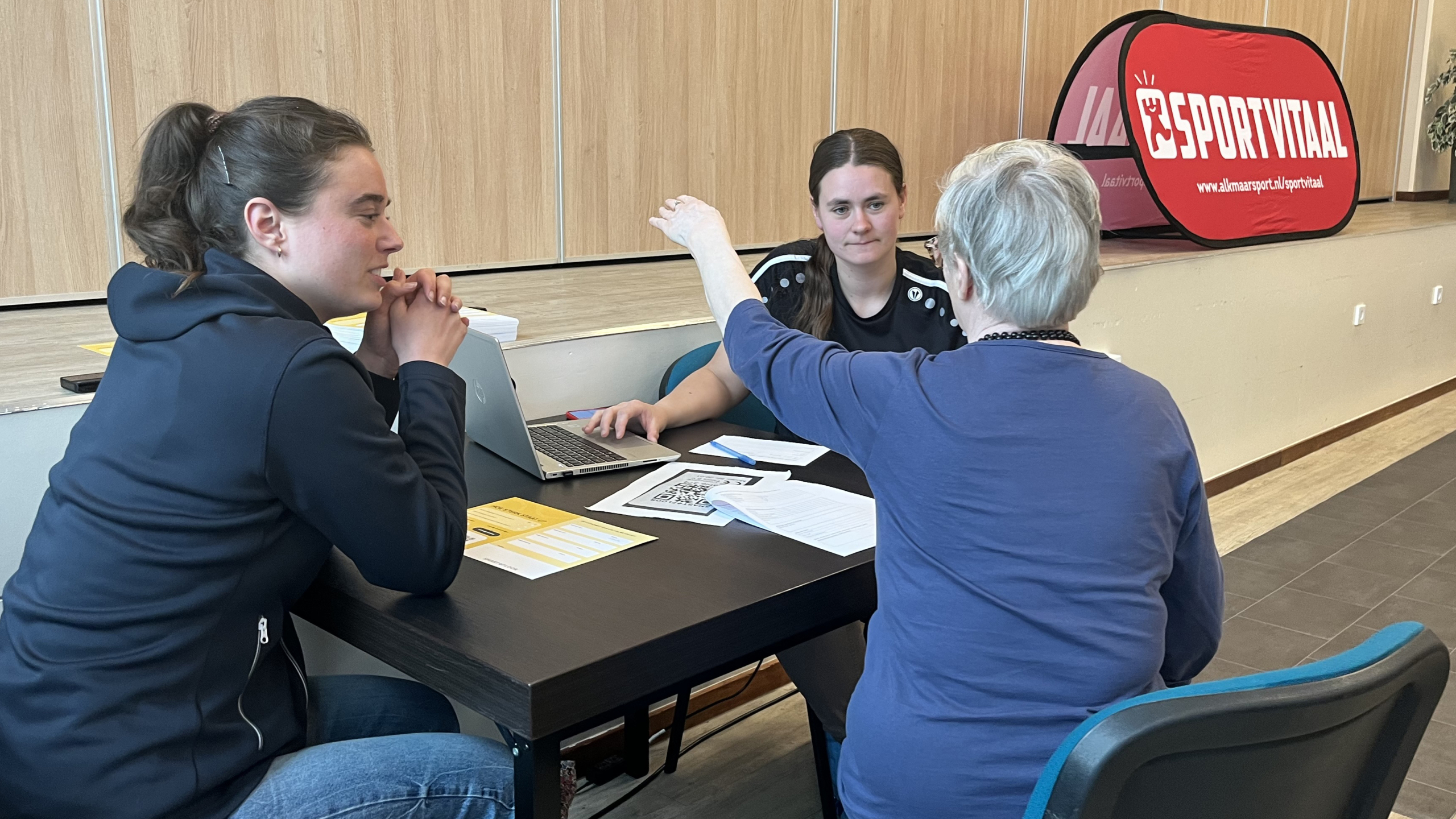 Drie vrouwen zitten rond een tafel met papieren en een laptop, in gesprek, met op de achtergrond een rode banner met 'SPORTVITAAL' erop.