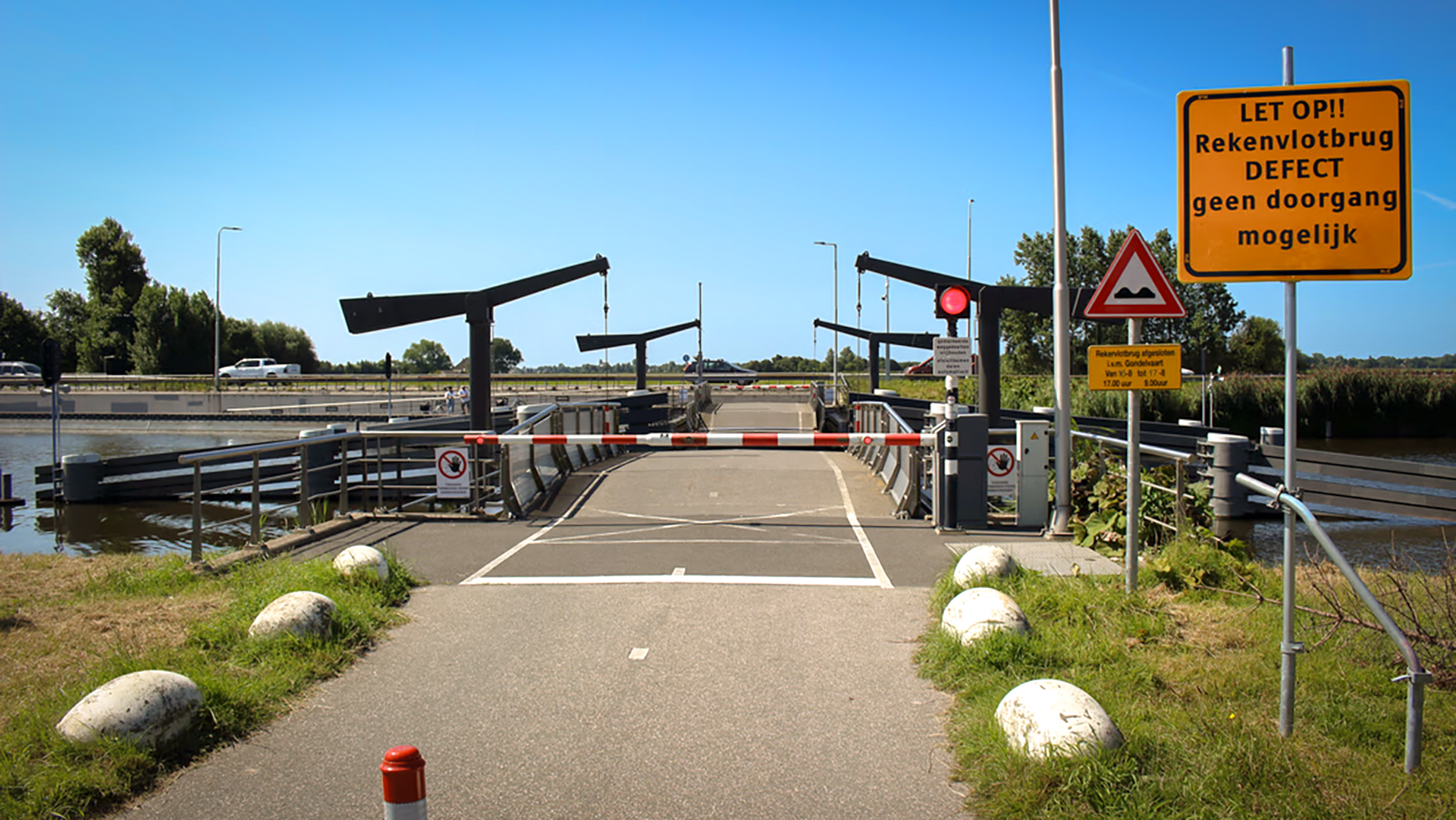 Een defecte brug met een gele waarschuwingsbord die vermeldt dat de Rekenvlotbrug niet toegankelijk is, terwijl er rode verkeerslichten en slagbomen zichtbaar zijn op een zonnige dag langs een kanaal, met gras en een weg eromheen.