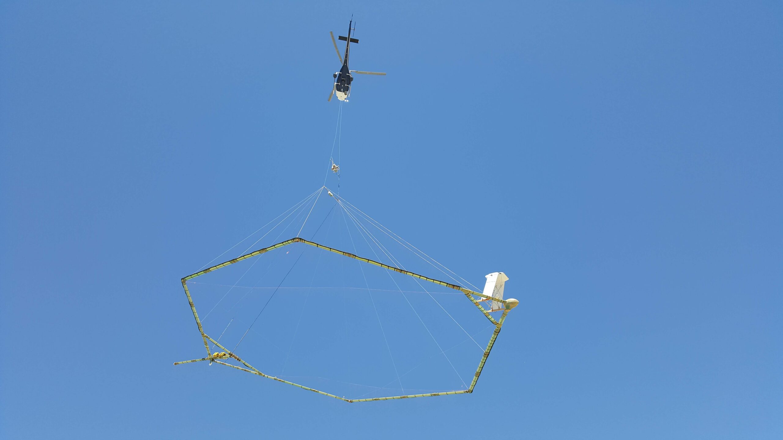Een helikopter vervoert een groot, zeshoekig geofysisch meetinstrument in de lucht tegen een heldere blauwe hemel.