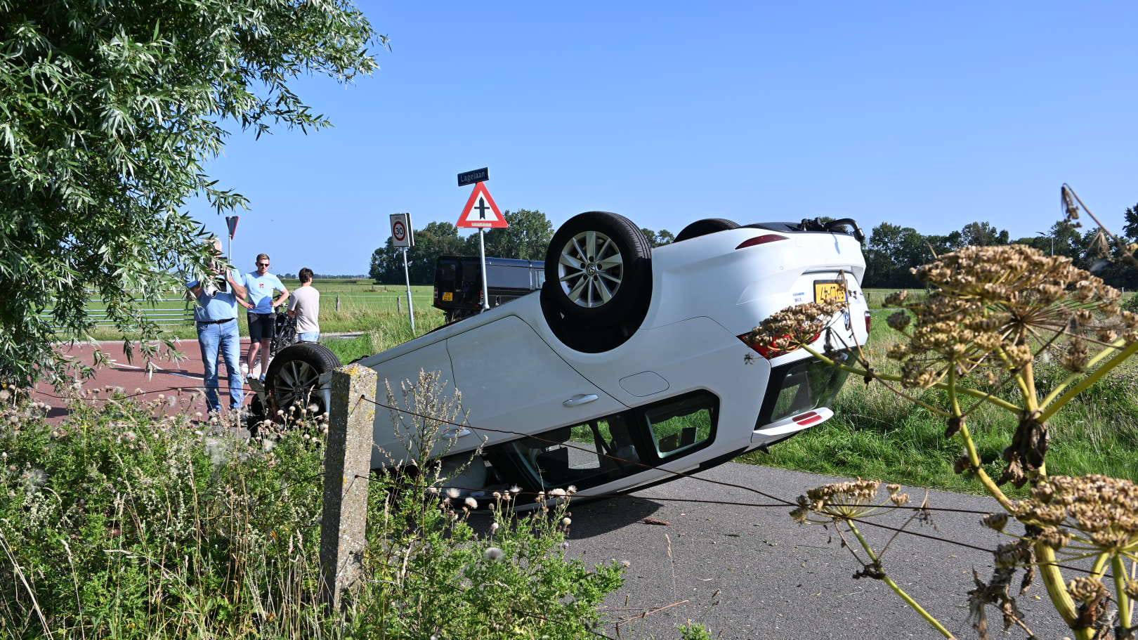Een witte auto ligt ondersteboven op een weg met een groepje mensen ernaast die de situatie bekijken, omringd door groen en een blauwe lucht.