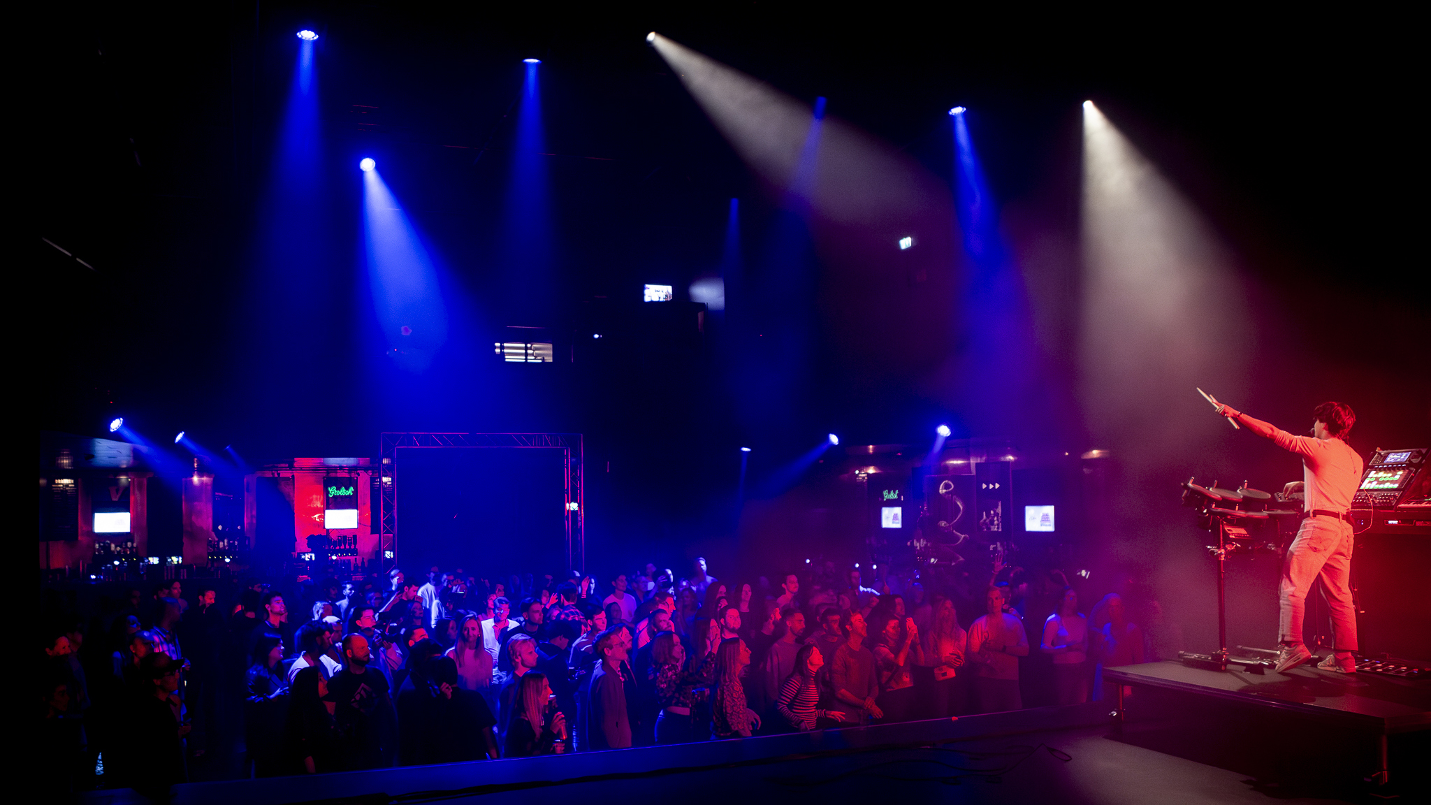 Een DJ staat op een verhoogd podium tijdens een concert met een menigte toeschouwers die voor het podium staan in een zaal, verlicht door kleurrijke spotlights.