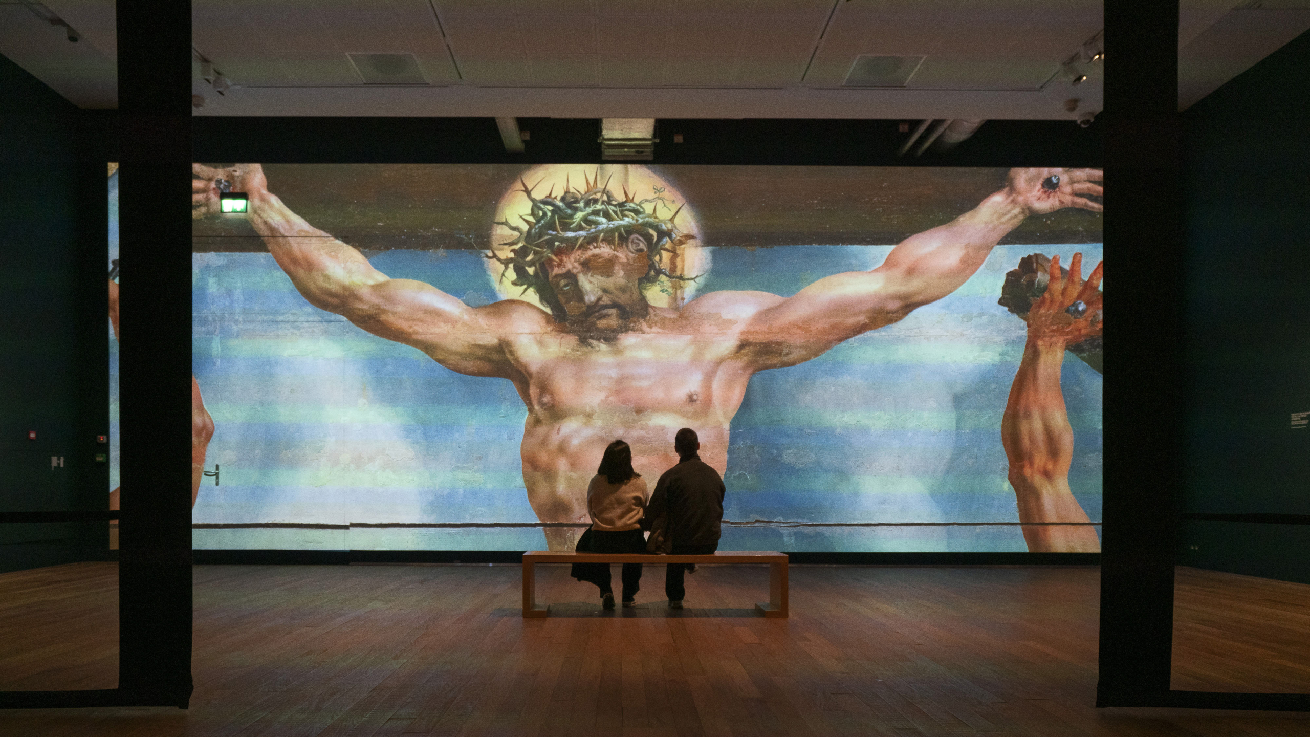 Twee mensen zitten op een bankje in een museum en kijken naar een grote projectie van een religieus kunstwerk met Jezus aan het kruis, gekroond met doornen.