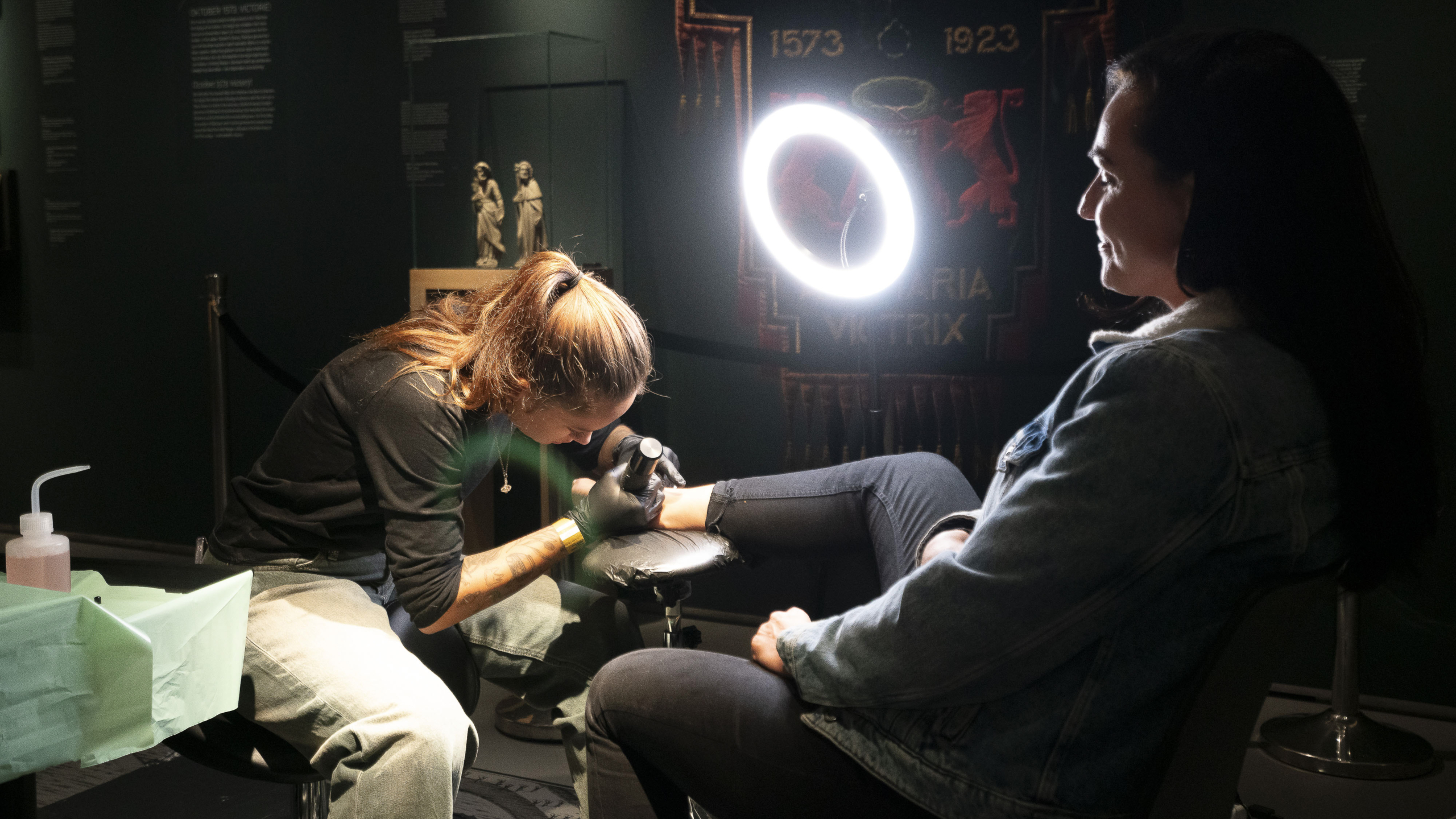 Een tatoeëerder bezig met het tatoeëren van een vrouw in een donkere kamer, met een heldere ringlamp die het gebied verlicht.