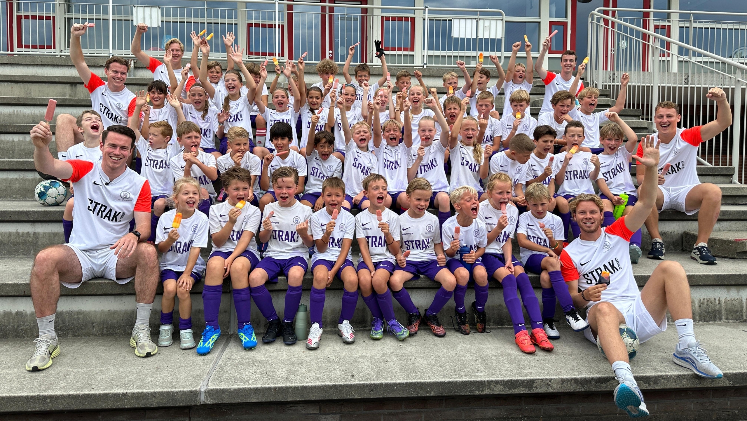 Een grote groep kinderen en volwassenen, gekleed in witte voetbalshirts met "STRAK!" erop, zitten enthousiast op tribunes. De kinderen houden ijsjes vast en lijken blij, terwijl de volwassenen naast hun poseren met opgeheven armen. Op de achtergrond is een gebouw met rode deuren en enkele borden te zien.