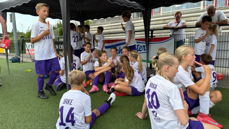 Kinderen in voetbalshirts zitten onder een zonnetent op kunstgras en eten waterijsjes tijdens een pauze in een sportevenement.
