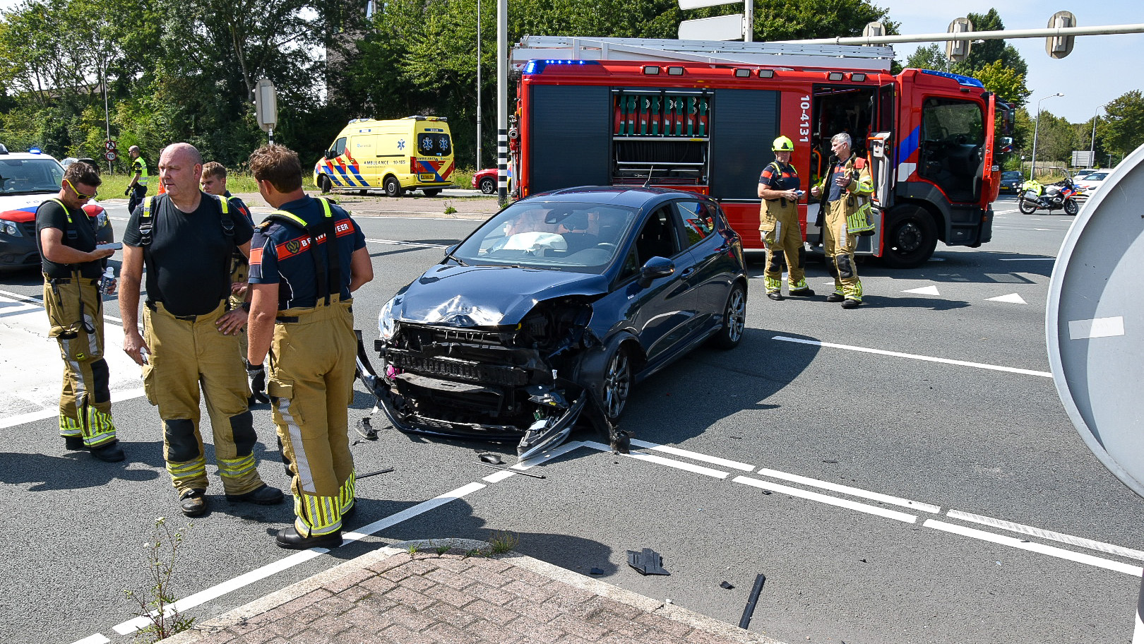 Een auto met schade aan de voorkant staat op een weg, omringd door brandweerlieden en een brandweerwagen. Op de achtergrond staat een ambulance geparkeerd. Het incident vindt plaats op een kruispunt met enkele verkeersborden en fietspaden zichtbaar.