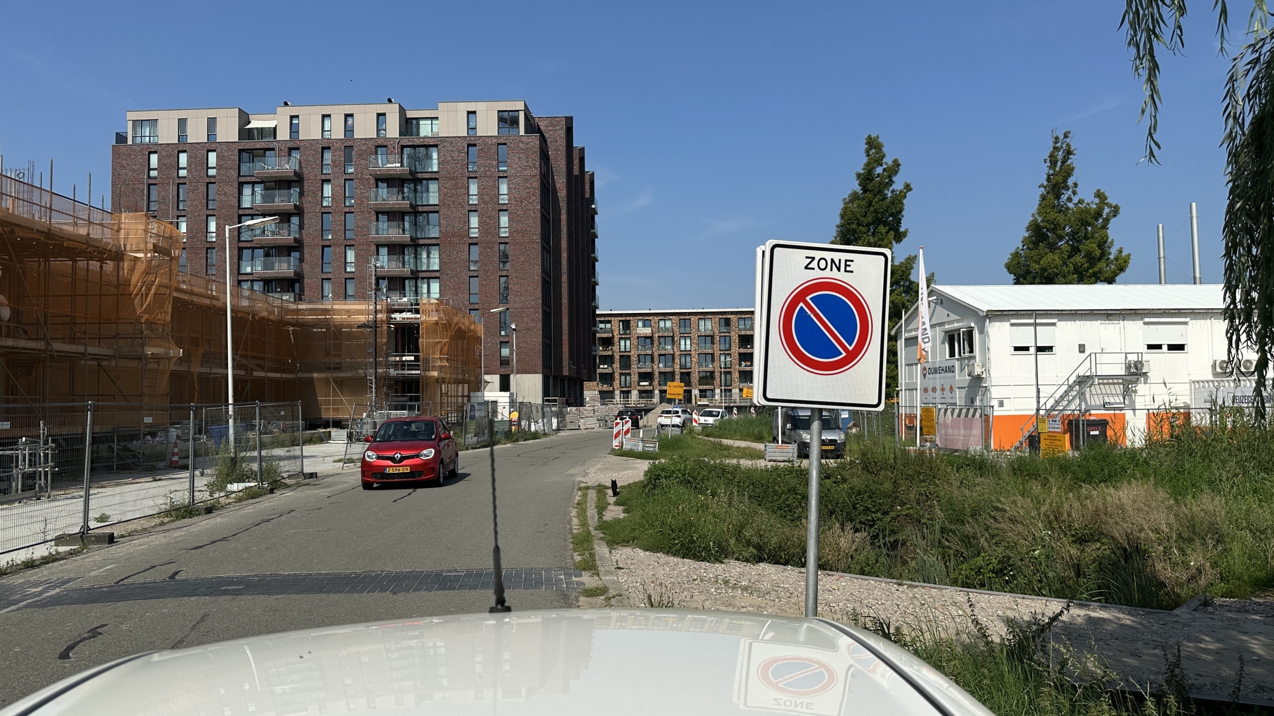 Gebouw in aanbouw met verschillende verdiepingen en een rode auto die door de straat rijdt, naast een verkeersbord dat een parkeerverbod aanduidt.