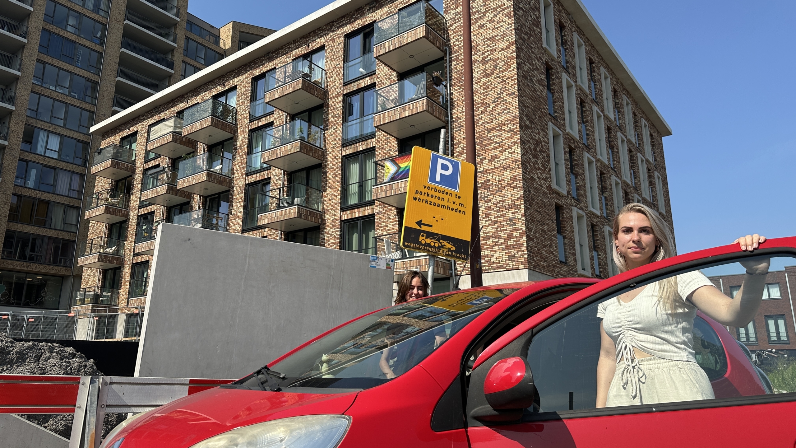 Een vrouw opent de deur van een rode auto voor een modern appartementencomplex met bakstenen gevel en balkons. Er is een verkeersbord dat parkeren verbiedt in verband met werkzaamheden. De lucht is helder en blauw.
