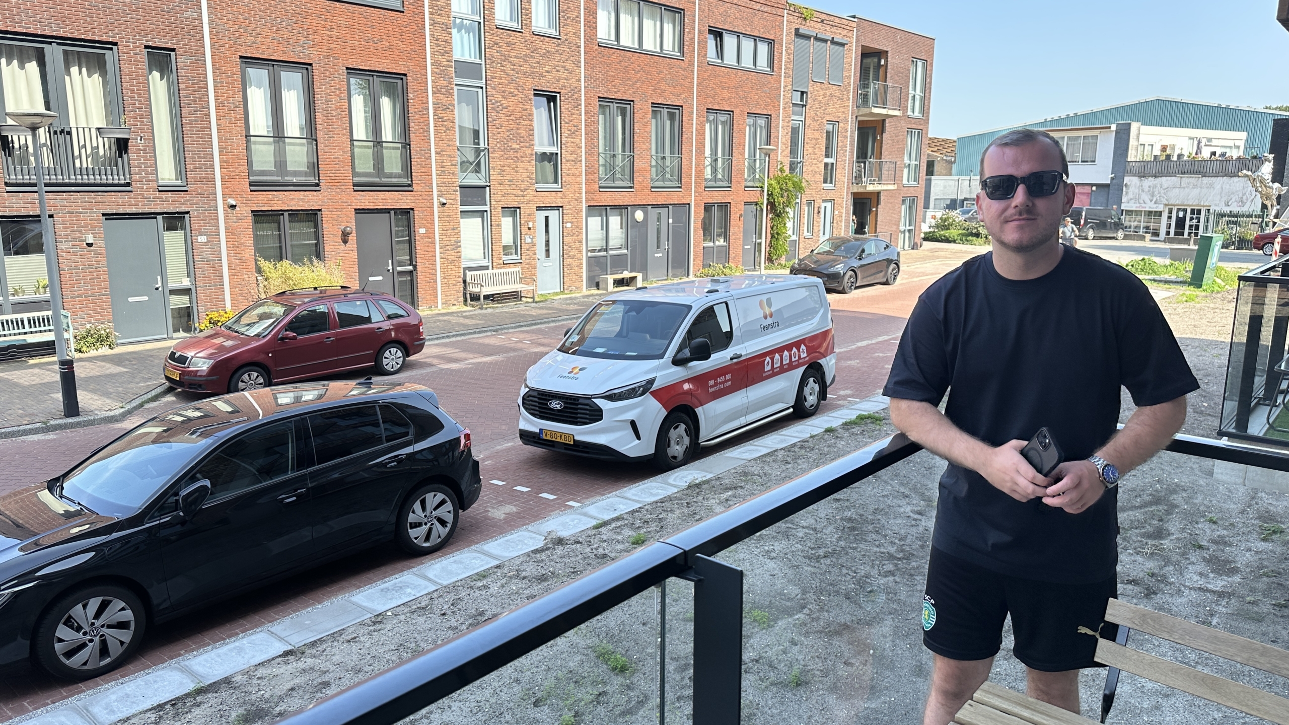 Een man met een zwarte zonnebril en een donker T-shirt staat op een balkon met een smartphone in zijn hand. Op de achtergrond zijn een straat met geparkeerde auto's en moderne bakstenen gebouwen zichtbaar.