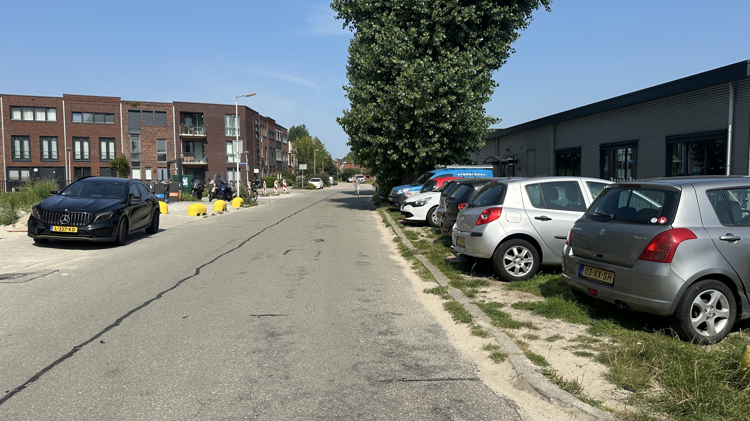 Straatbeeld met geparkeerde auto's langs de weg, waaronder een zwarte auto en meerdere zilverkleurige voertuigen, naast een gebouw met bakstenen gevels en enkele bomen langs de stoep.