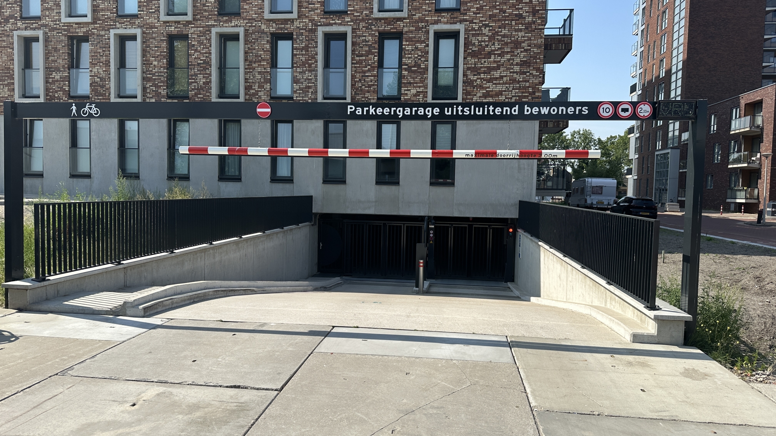 Toegang tot een ondergrondse parkeergarage voor uitsluitend bewoners, met een slagboom en verkeersbord met hoogtebeperking, omringd door een moderne bakstenen flat.