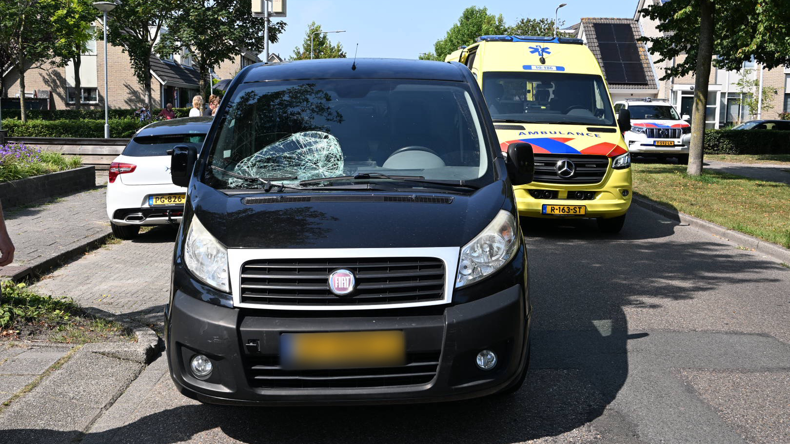 Een zwarte Fiat bestelwagen met een verbrijzeld voorruit staat op straat, met daarachter een gele ambulance en een witte auto in een woonwijk.