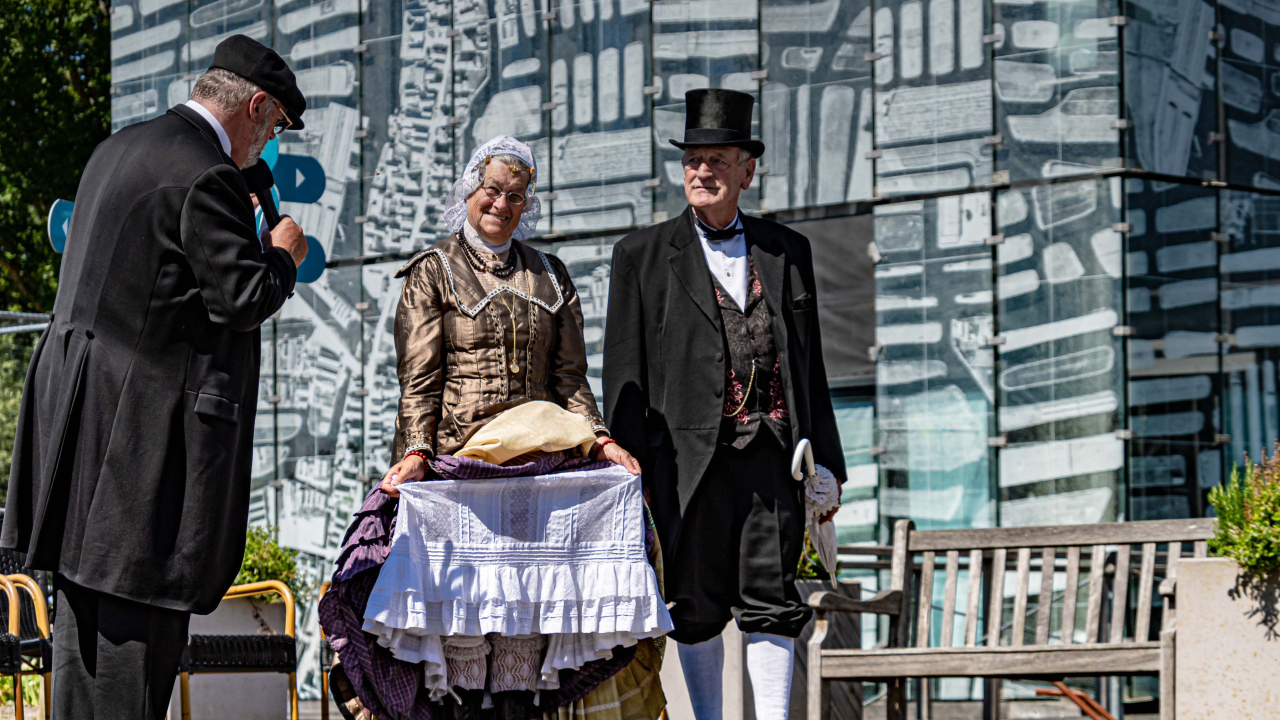 Twee mensen in historische kleding poseren op een podium met een man die een microfoon vasthoudt, tegen een achtergrond van een gebouw met glasgevels.