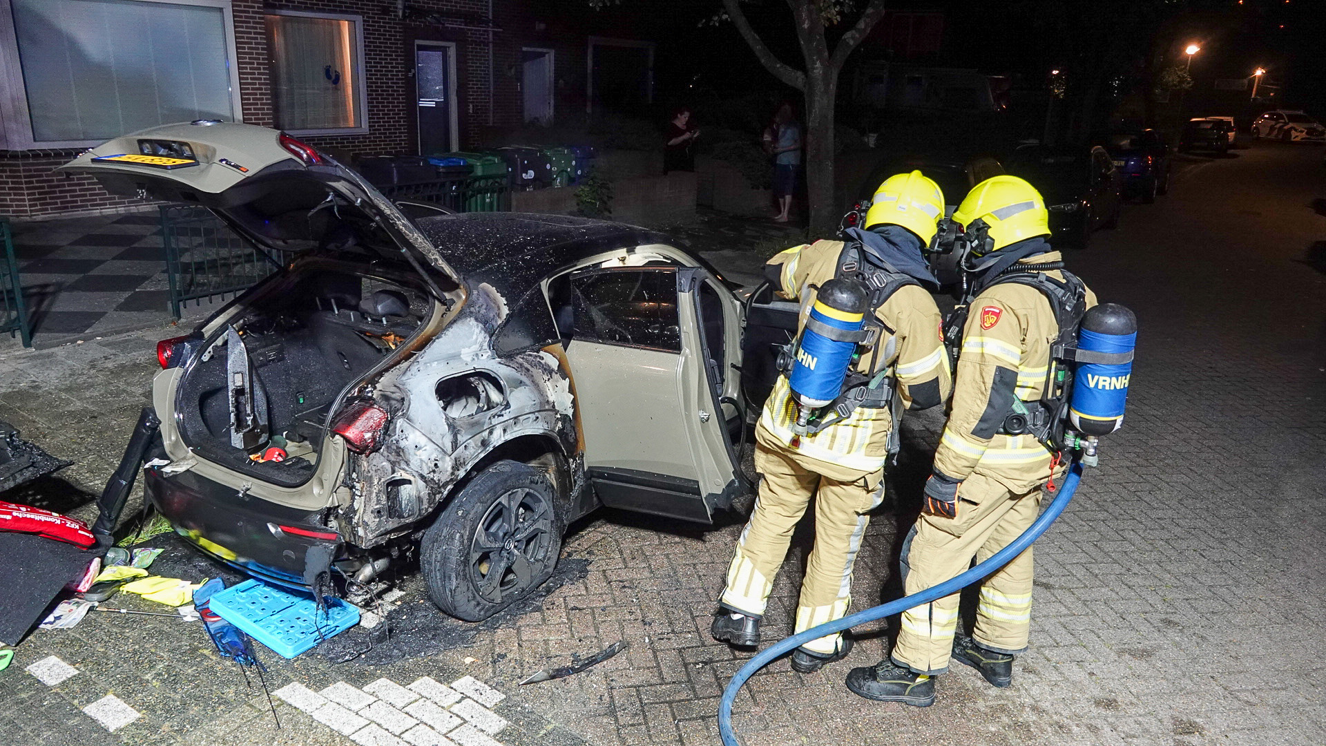 Twee brandweerlieden in uniform inspecteren een uitgebrande auto met open kofferruimte, staande op een straat bij nacht.