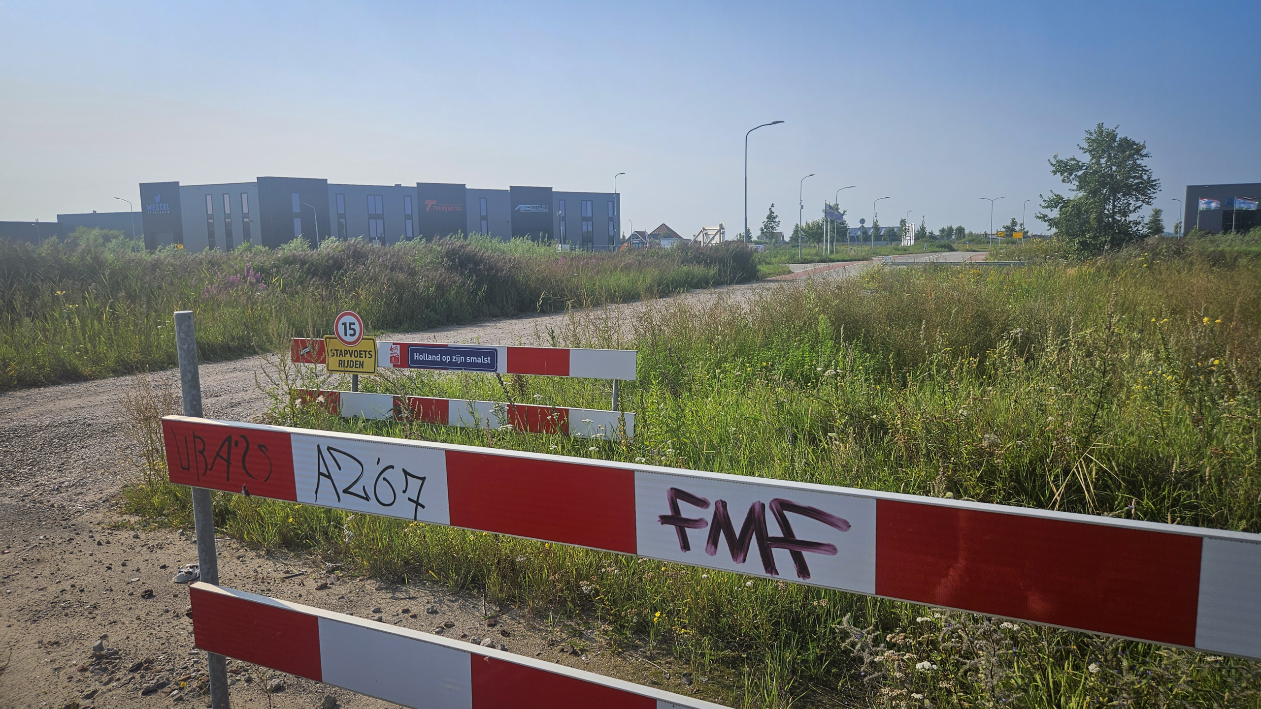 Een bouwterrein met borden en rode-witte afzettingen, waarop teksten zoals "Holland op zijn smalst" en graffiti te zien zijn. Op de achtergrond zijn industriële gebouwen en straatlantaarns zichtbaar, omgeven door hoog gras.
