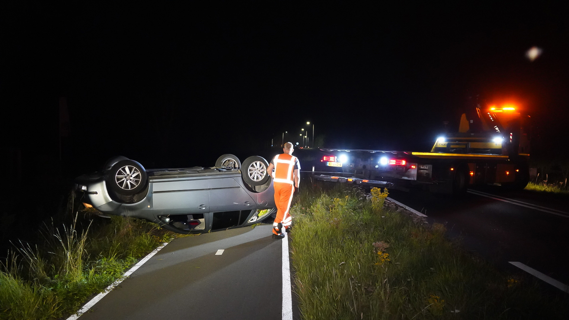 Een gekantelde auto ligt op zijn dak op een weg 's nachts, terwijl een persoon in oranje veiligheidskleding naar de scene loopt. In de achtergrond staat een takelwagen met geopende lichten.