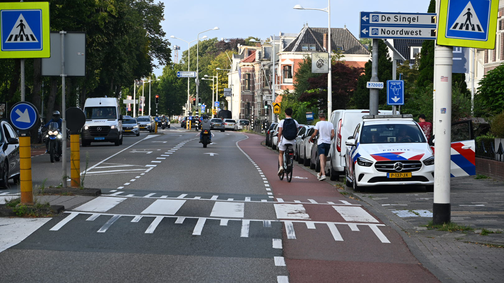 Straatbeeld met een fietspad en een voetgangerspad langs een drukke weg, waar een politieauto en andere voertuigen geparkeerd staan; verkeersborden wijzen de richting naar "De Singel" en "Noordwest."