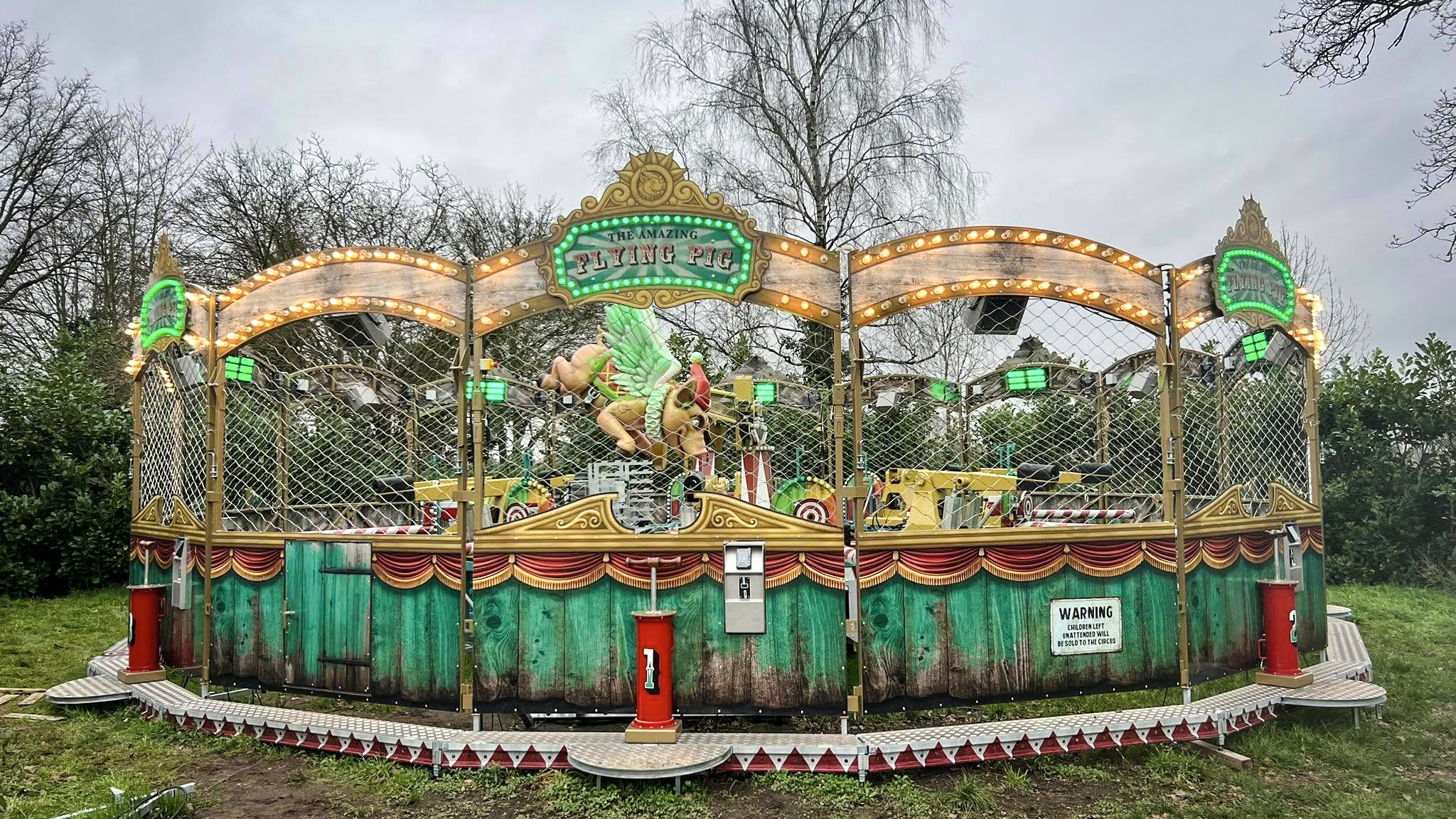 Een kleurrijke kermisattractie genaamd 'The Amazing Flying Pig' met verlichte borden en een decoratief ontwerp, omgeven door bomen op een bewolkte dag.
