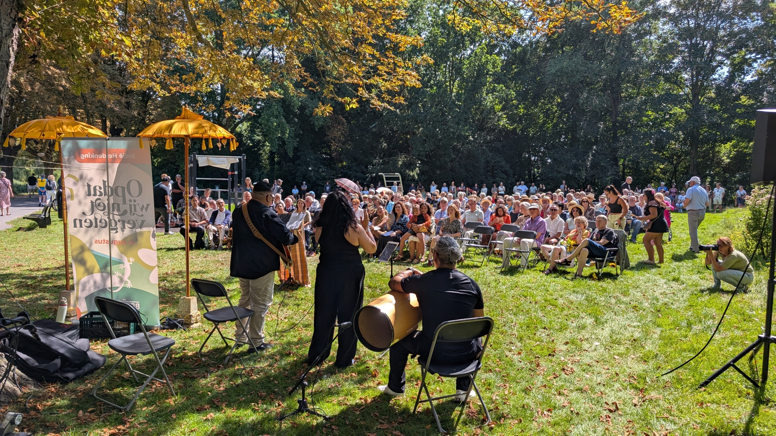 Muzikanten spelen voor een groot publiek op een zonnige dag in een park, met een banner en gele parasols op de voorgrond.