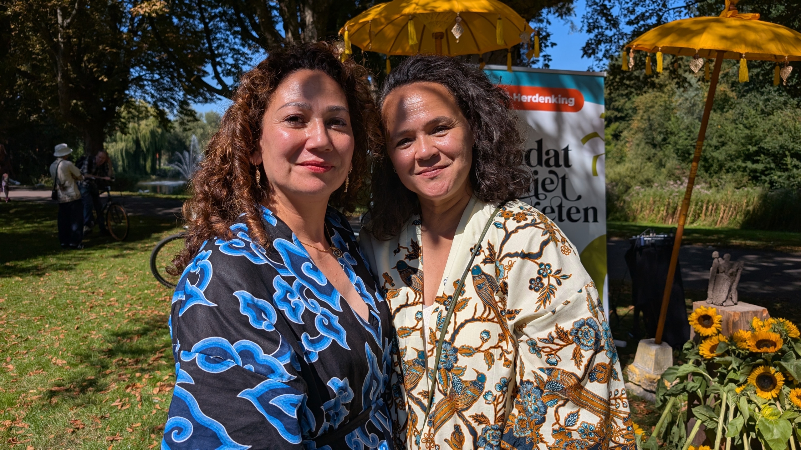 Twee vrouwen poseren samen buiten, omringd door een parkachtige omgeving met zonnebloemen en gele paraplu's.