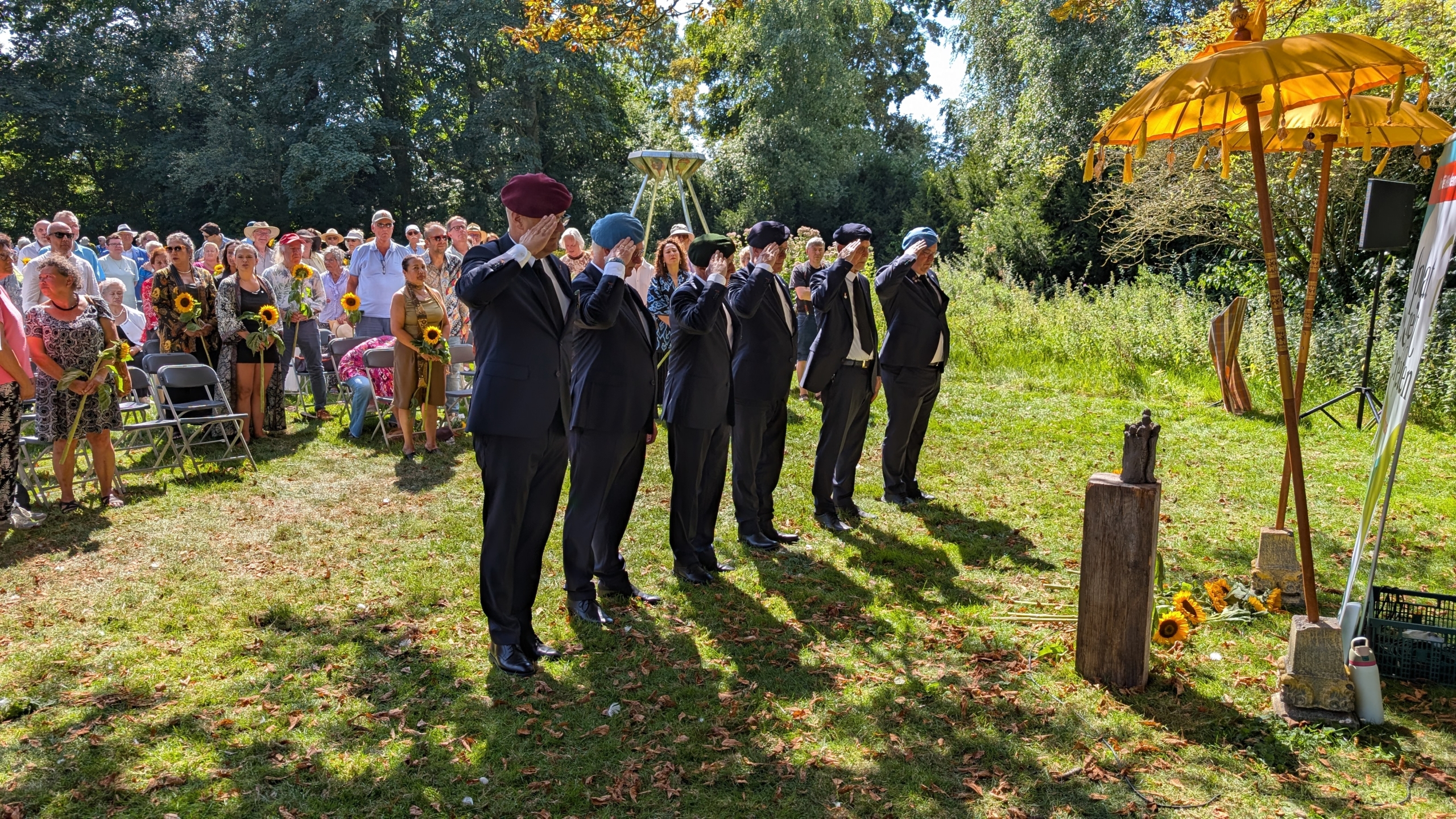 Een groep mensen, waarvan enkelen in uniform met baret, staan gesaluuterd op een ceremonie in een parkachtige omgeving, omringd door toeschouwers met zonnebloemen in hun handen, en onder een gele parasol.
