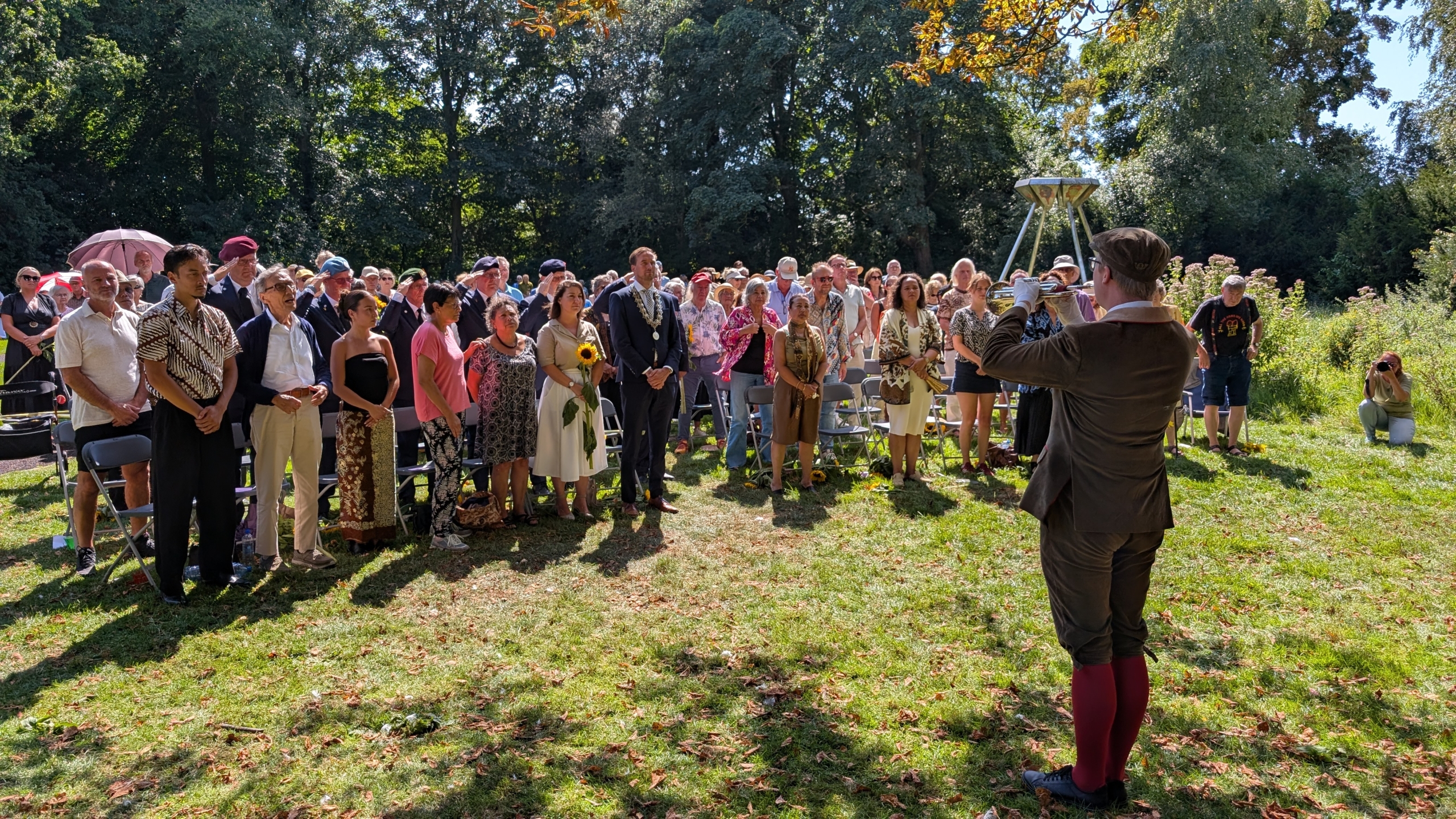 Een groep mensen staat buiten in een parkachtige omgeving, luisterend naar een persoon die een instrument bespeelt. De mensen lijken deel te nemen aan een ceremonie of herdenking. Enkele aanwezigen dragen formele kleding en sommigen houden bloemen vast. In de achtergrond zijn bomen te zien.