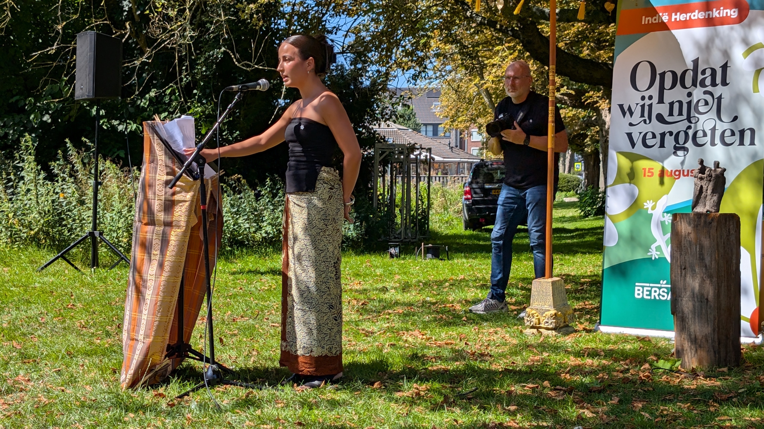 Vrouw spreekt in een microfoon buiten bij een herdenkingsceremonie, naast een groot bord met de tekst 'Indië Herdenking - Opdat wij niet vergeten - 15 augustus'.