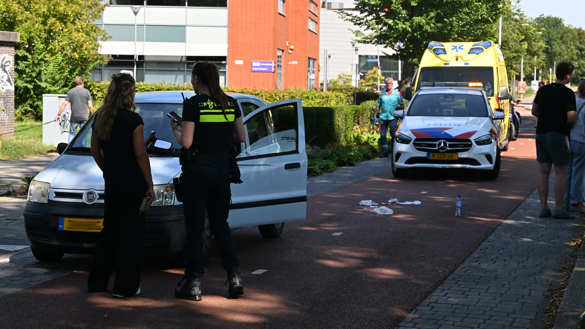 Een politieagent en een vrouw praten naast een witte auto met geopende deur, terwijl een politiewagen en ambulance op de achtergrond staan geparkeerd op een straat naast een gebouw.